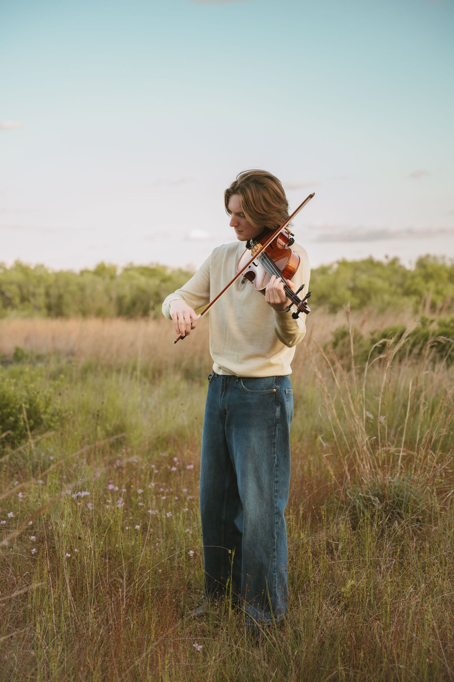 High school senior playing the violin against blue sky at graduation portrait session at Lake Runnymede Conservation Area St. Cloud Florida