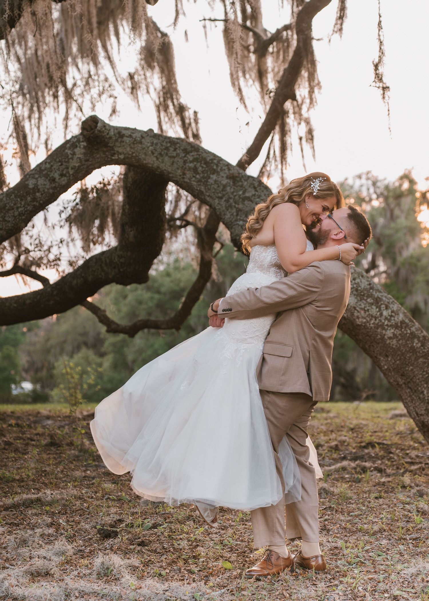 Romantic sunset portrait of the groom picking up his bride beneath the sprawling live oak trees at Moss Estates wedding venue in Orlando