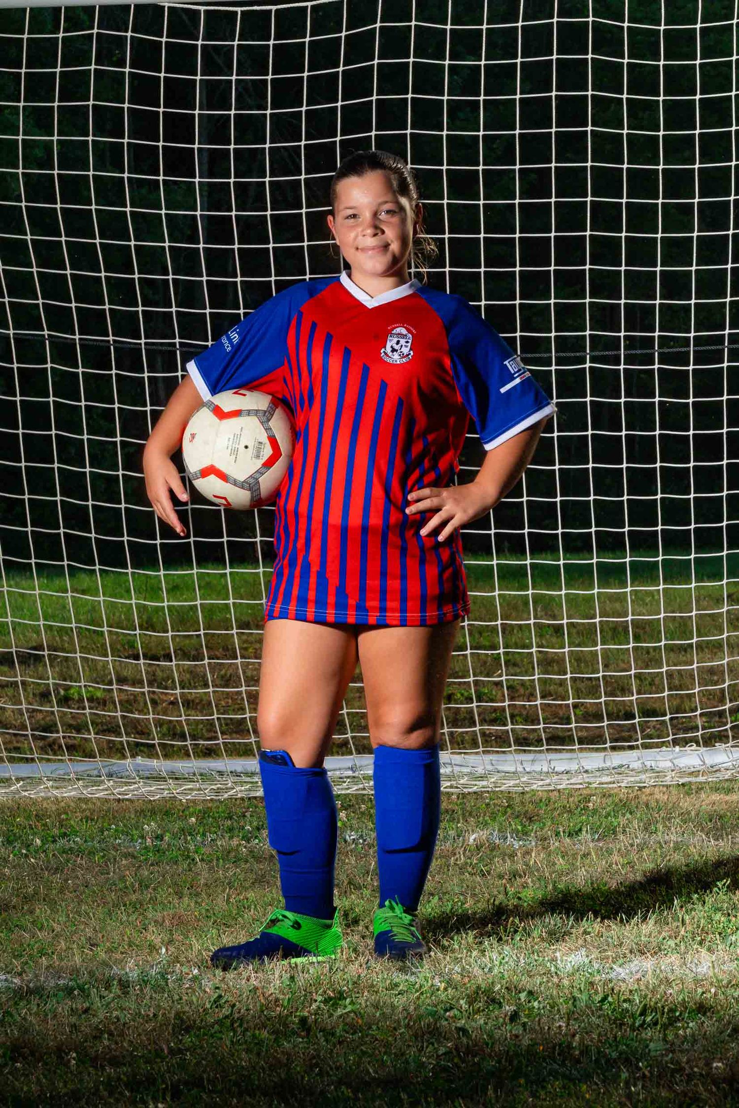 Night soccer team photography showing an individual female player standing on a turf field holding a soccer ball.
