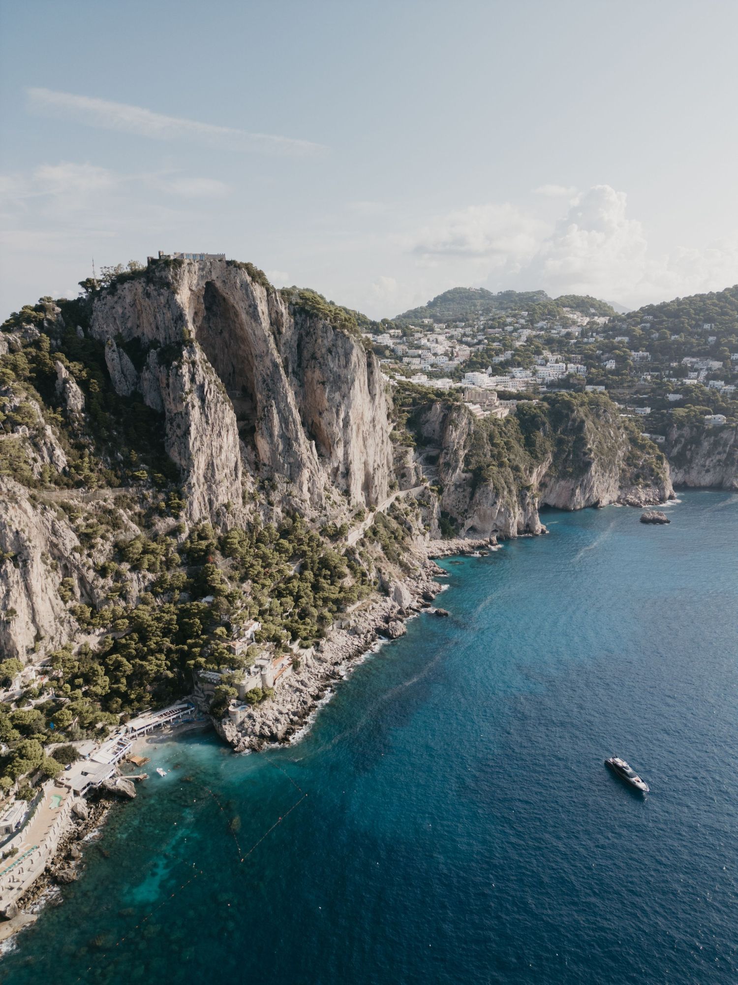 Dramatic coastal cliffs of Capri Island rise above turquoise Mediterranean waters with luxury yachts sailing below.