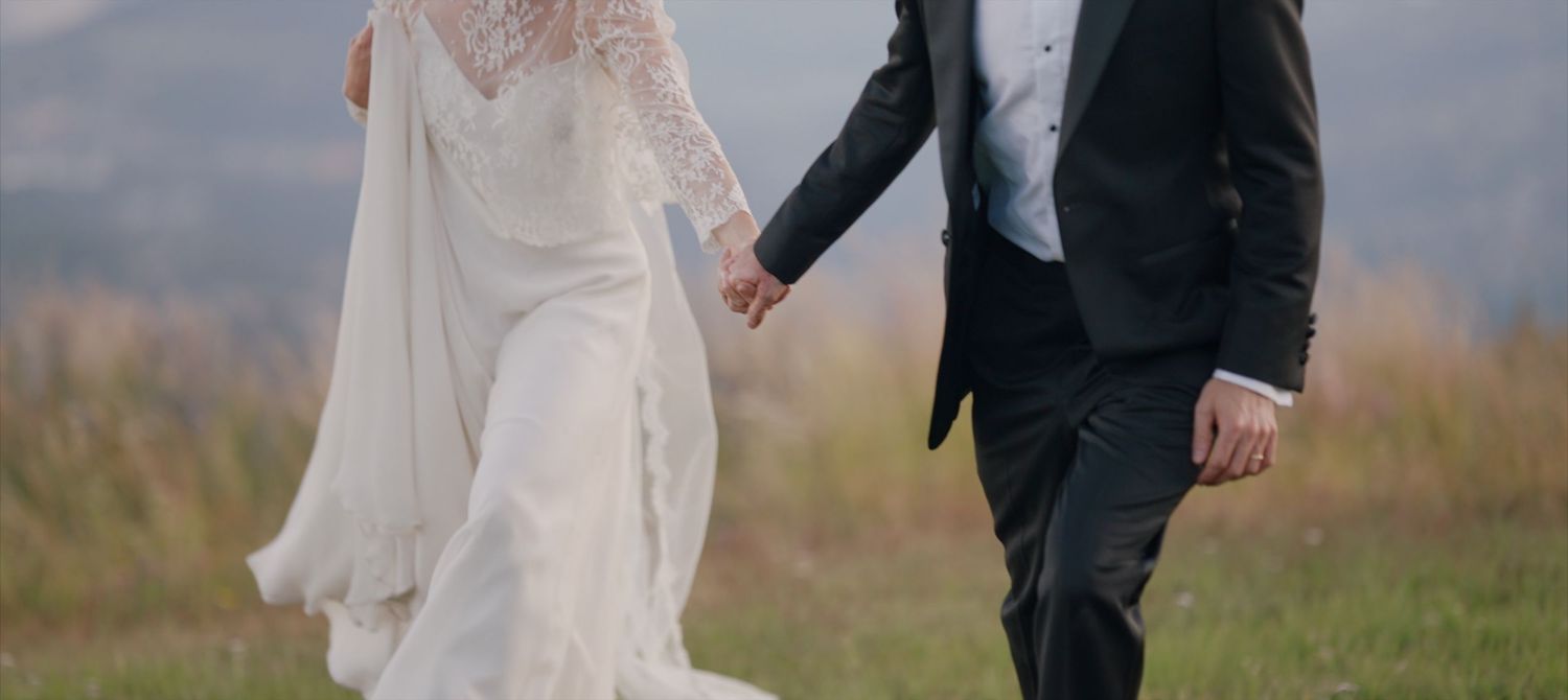A couple in wedding attire walks hand in hand through a grassy meadow on their special day.
