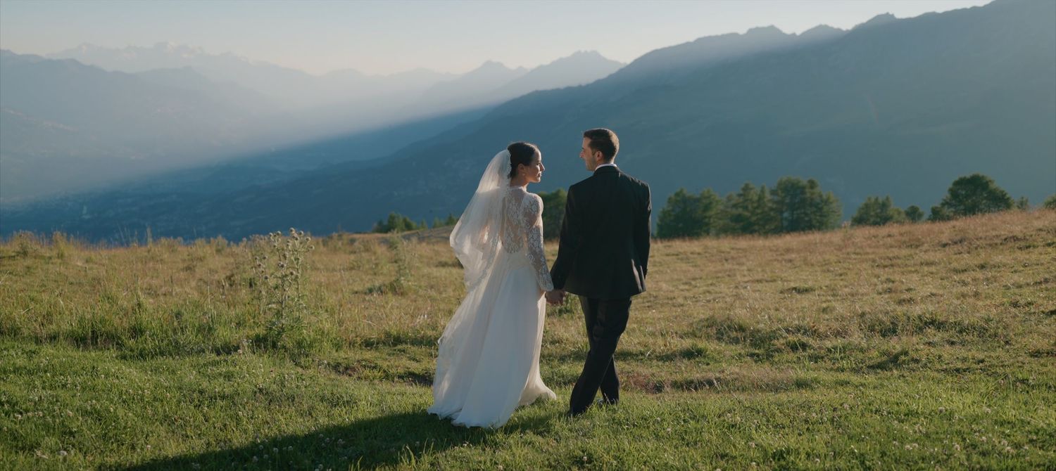A couple walks along a scenic Swiss mountain ridge at sunset with dramatic mountain peaks in the background.