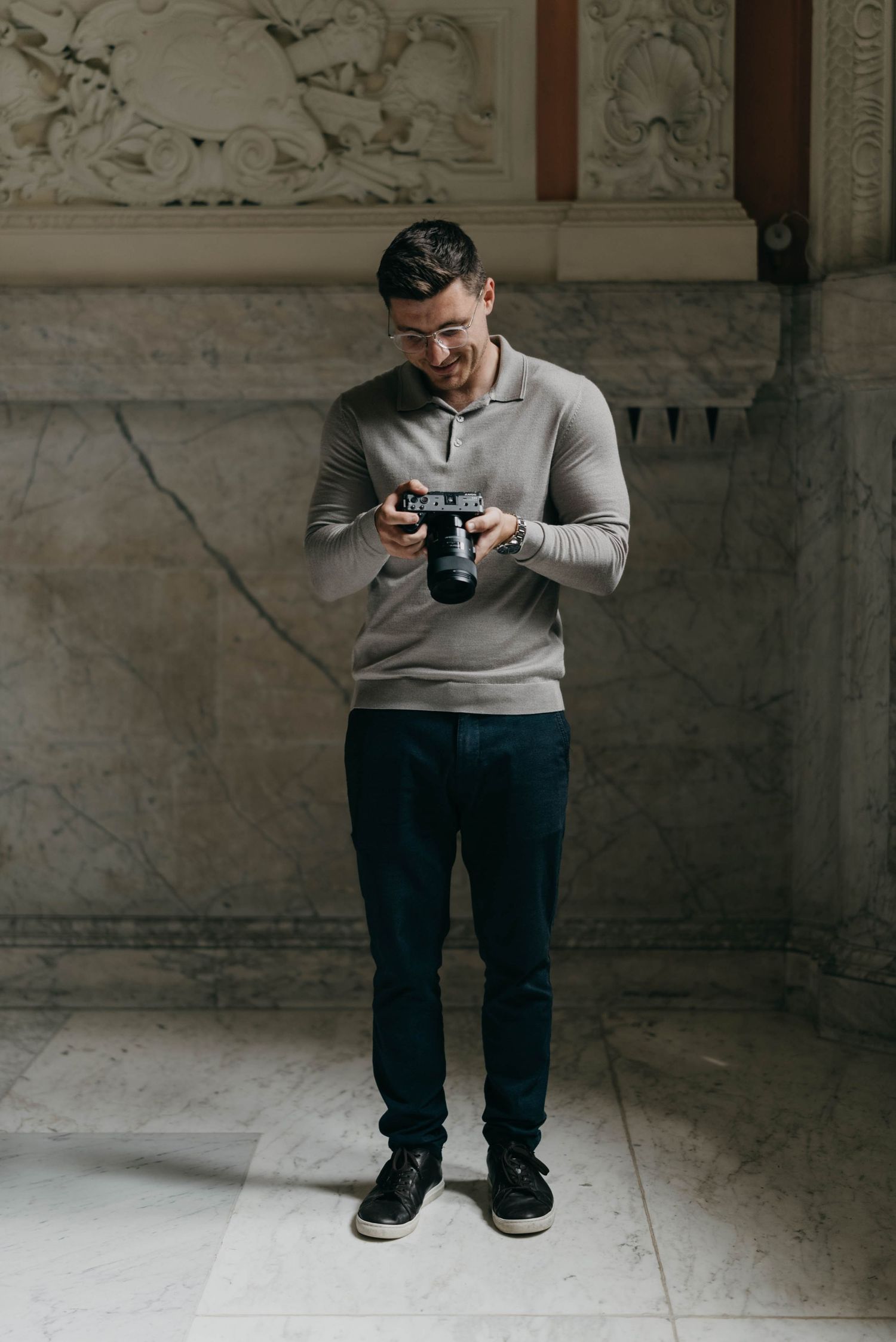 Jake Burgess in grey sweater and dark pants looking down at digital camera in a stone-walled interior space.