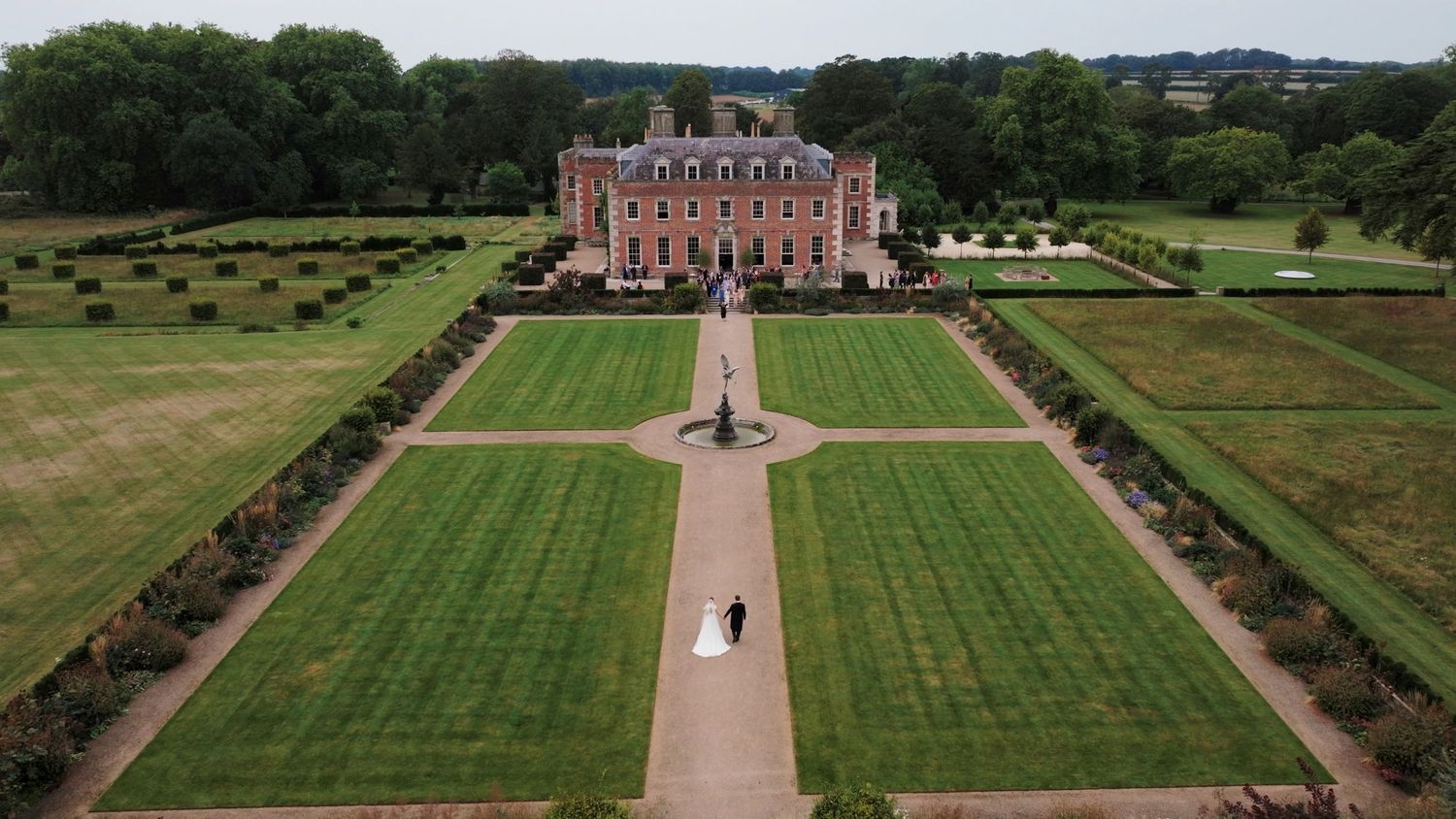 A panoramic view of a grand English estate with manicured lawns, formal gardens and a historic brick mansion.