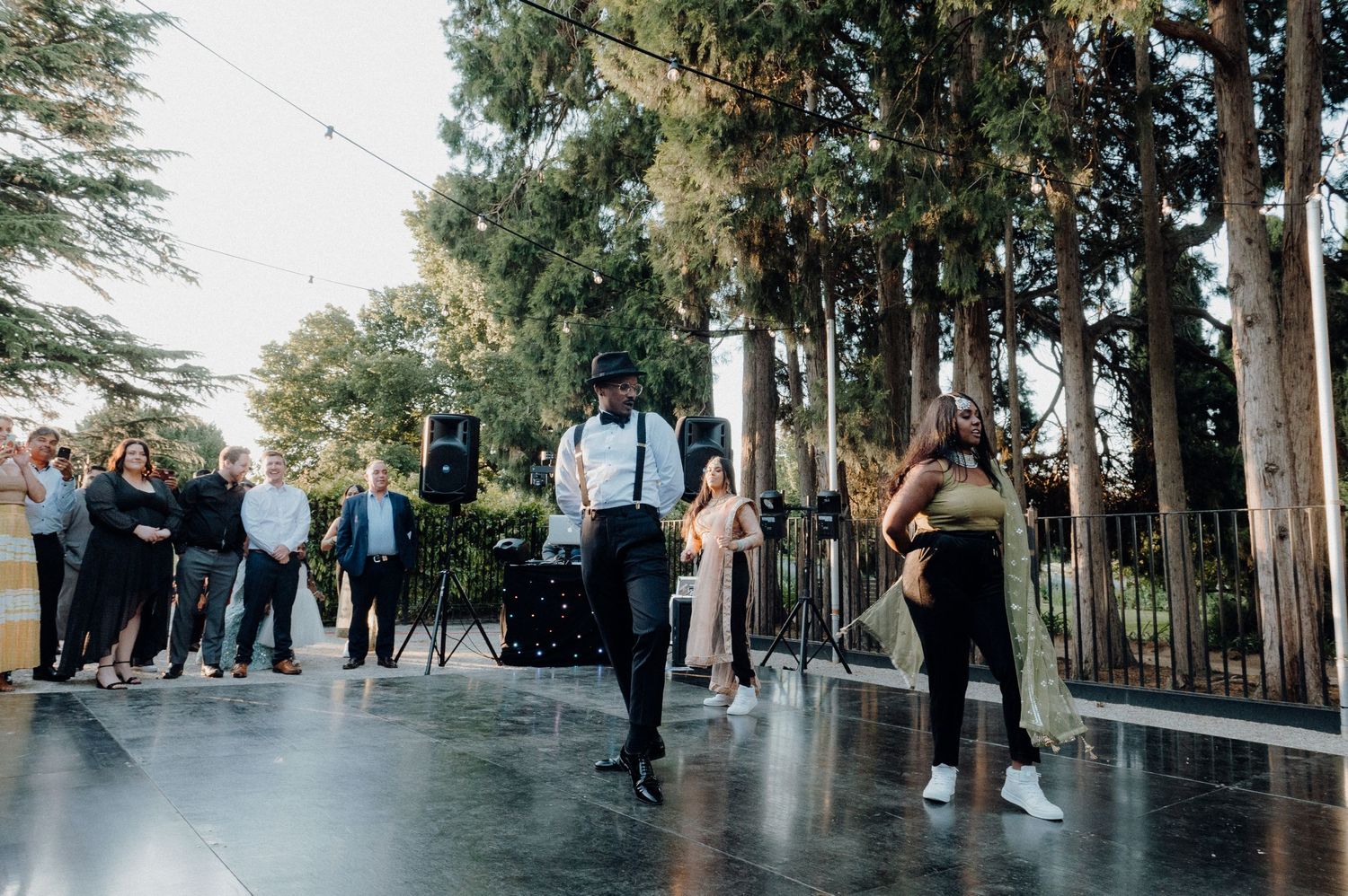 Wedding party dancing outdoors on a covered dance floor surrounded by tall trees.