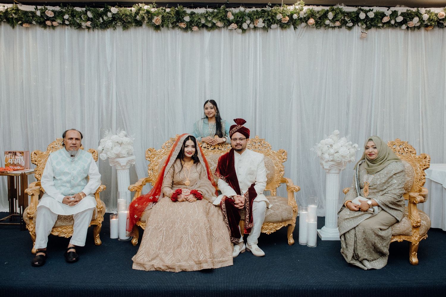 Wedding party seated on elegant chairs with floral decor and white draping.