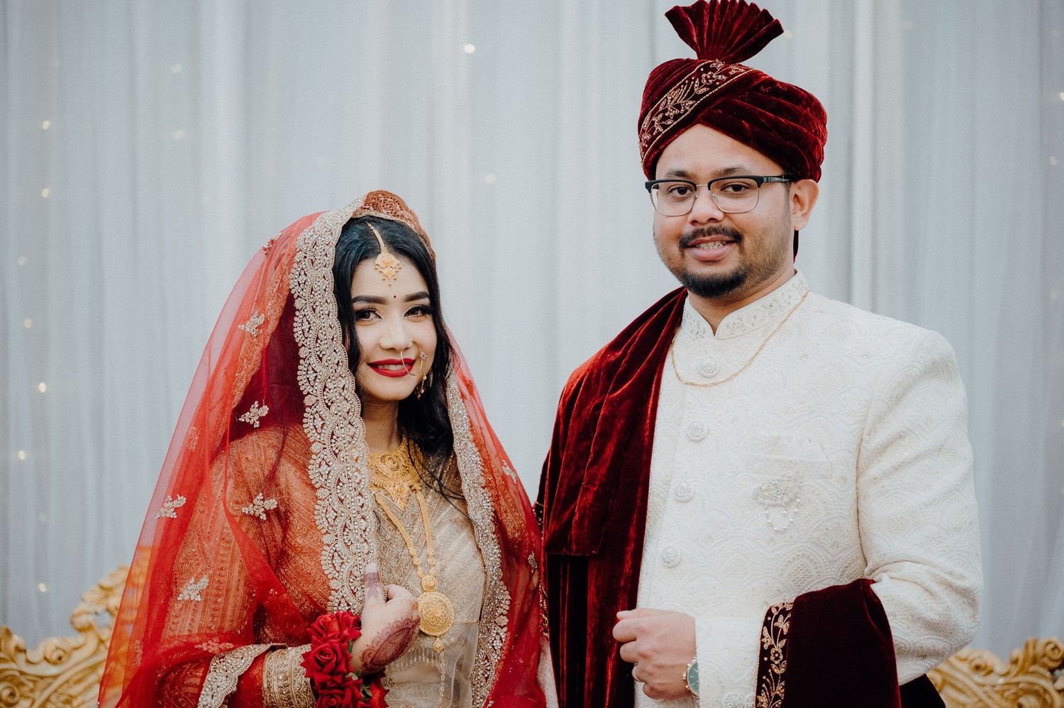 Newlyweds pose together in traditional South Asian wedding attire against a light backdrop.