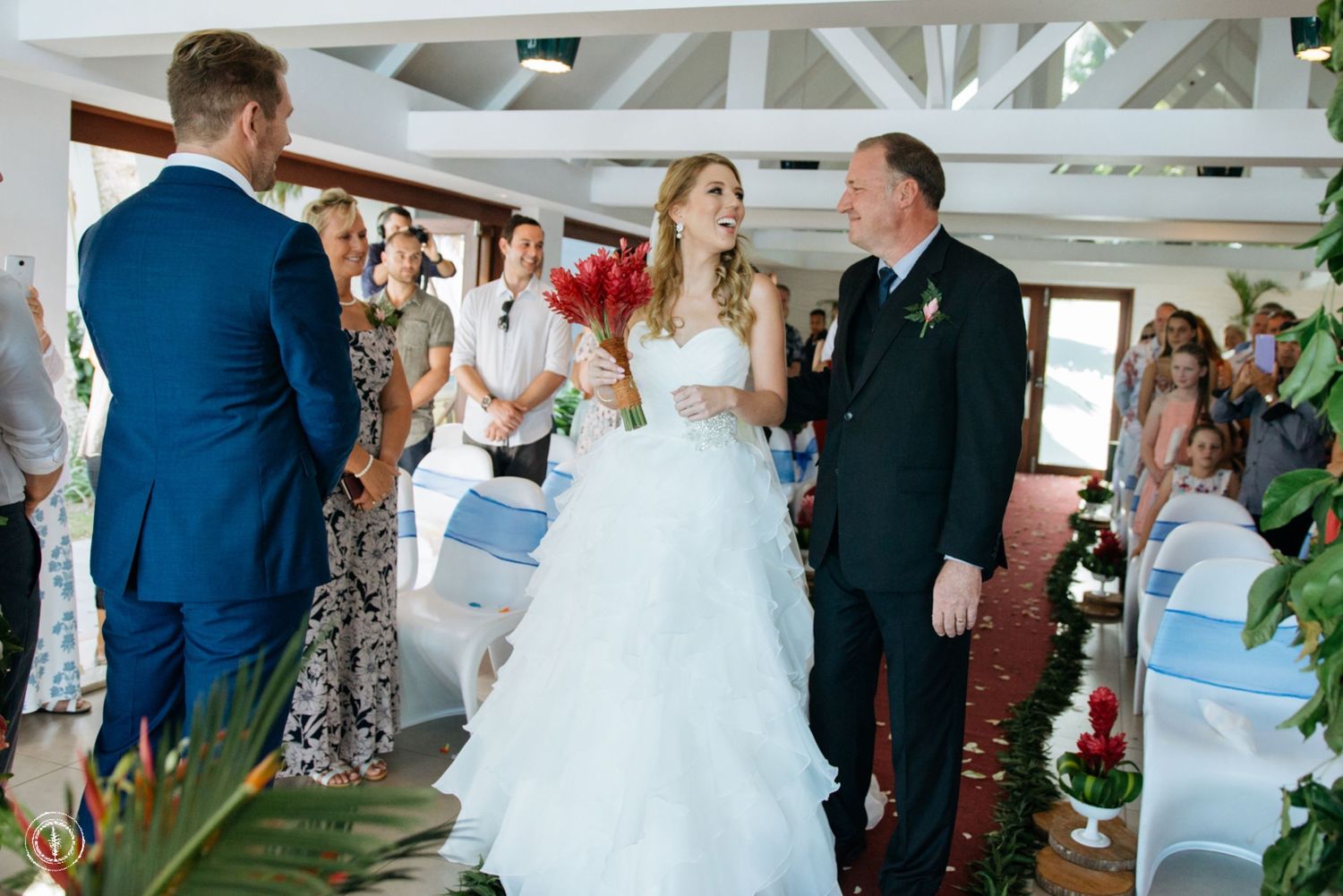 Wedding ceremony takes place under white beams with guests seated in chairs during an indoor celebration.