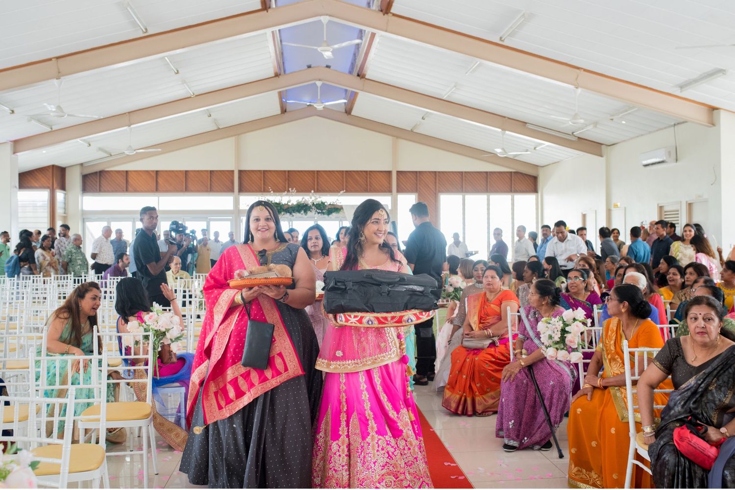 Wedding guests in colorful traditional attire gather in a bright reception hall with white chairs.