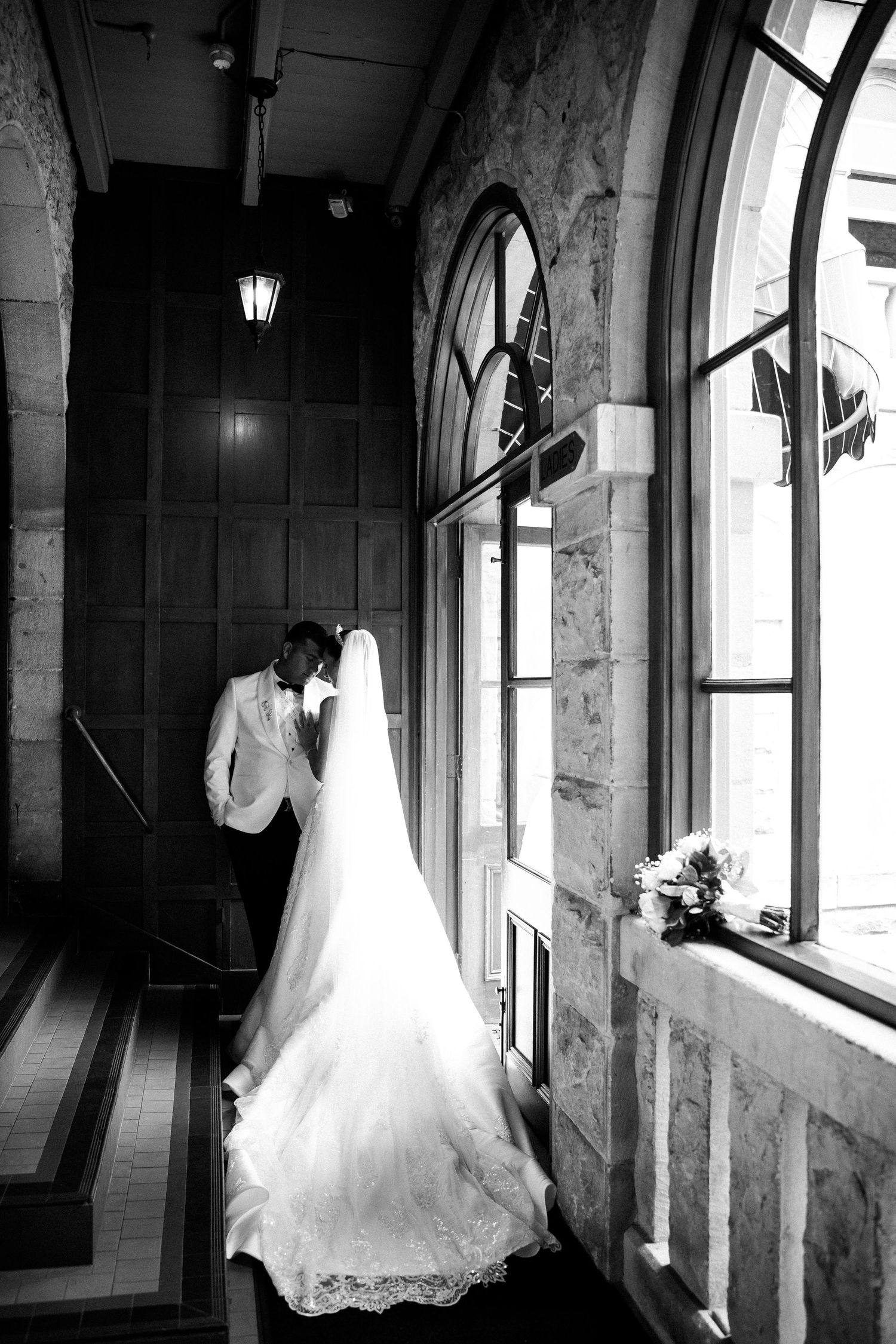 Bride and groom share intimate moment in black and white church corridor with arched windows.