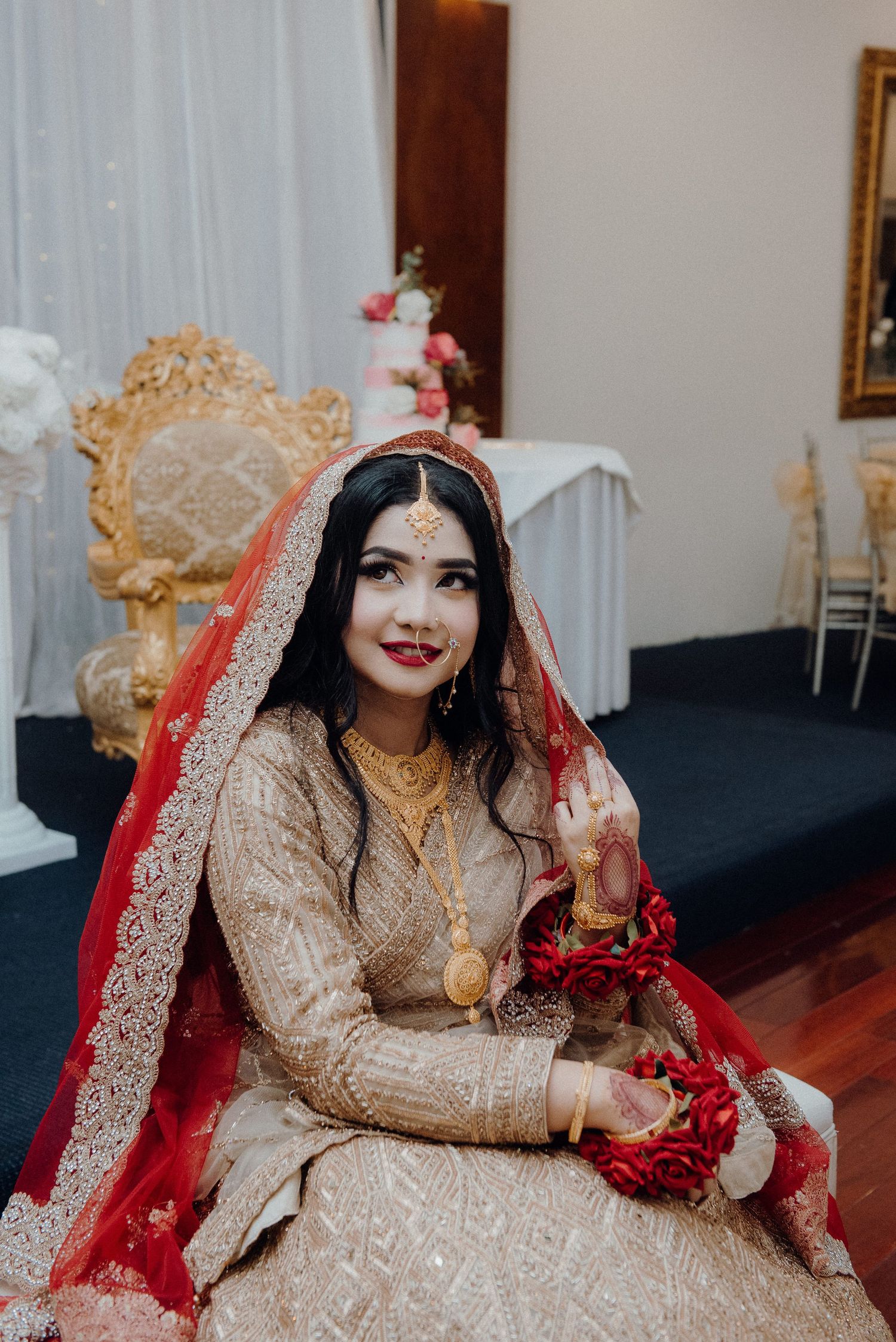 A bride in a red and gold lehenga poses elegantly on an ornate golden chair.