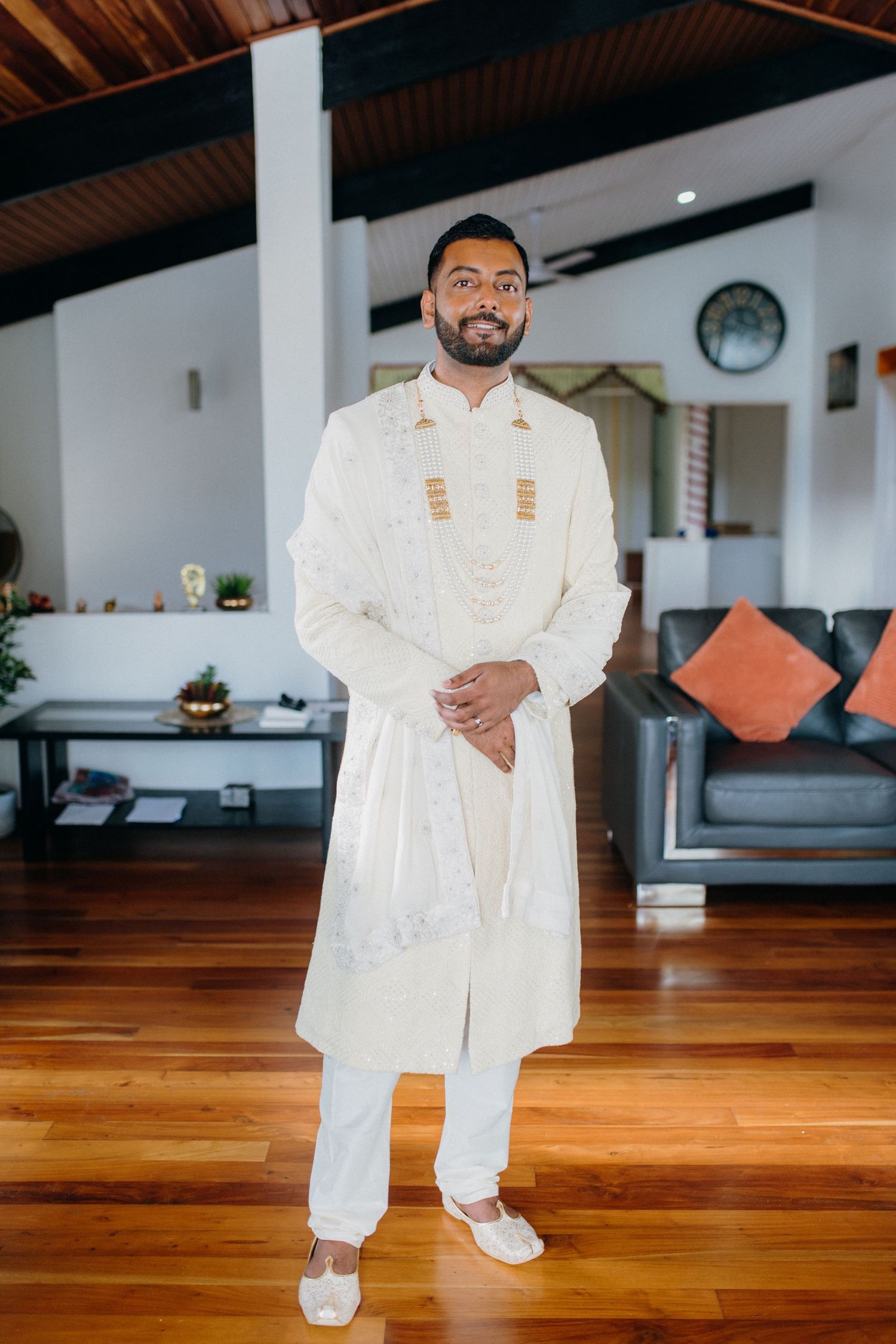 Person wearing a detailed white traditional Indian sherwani poses in an elegant living room setting.