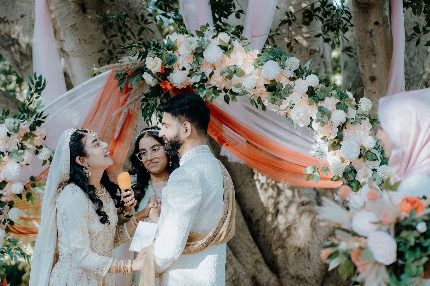 A couple shares a joyful moment under a floral arch decorated with orange and white flowers.