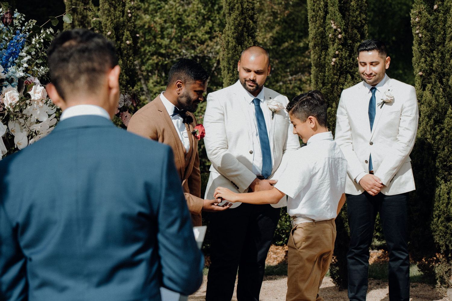 Wedding ceremony moment with attendees gathered under cypress trees and floral decorations.