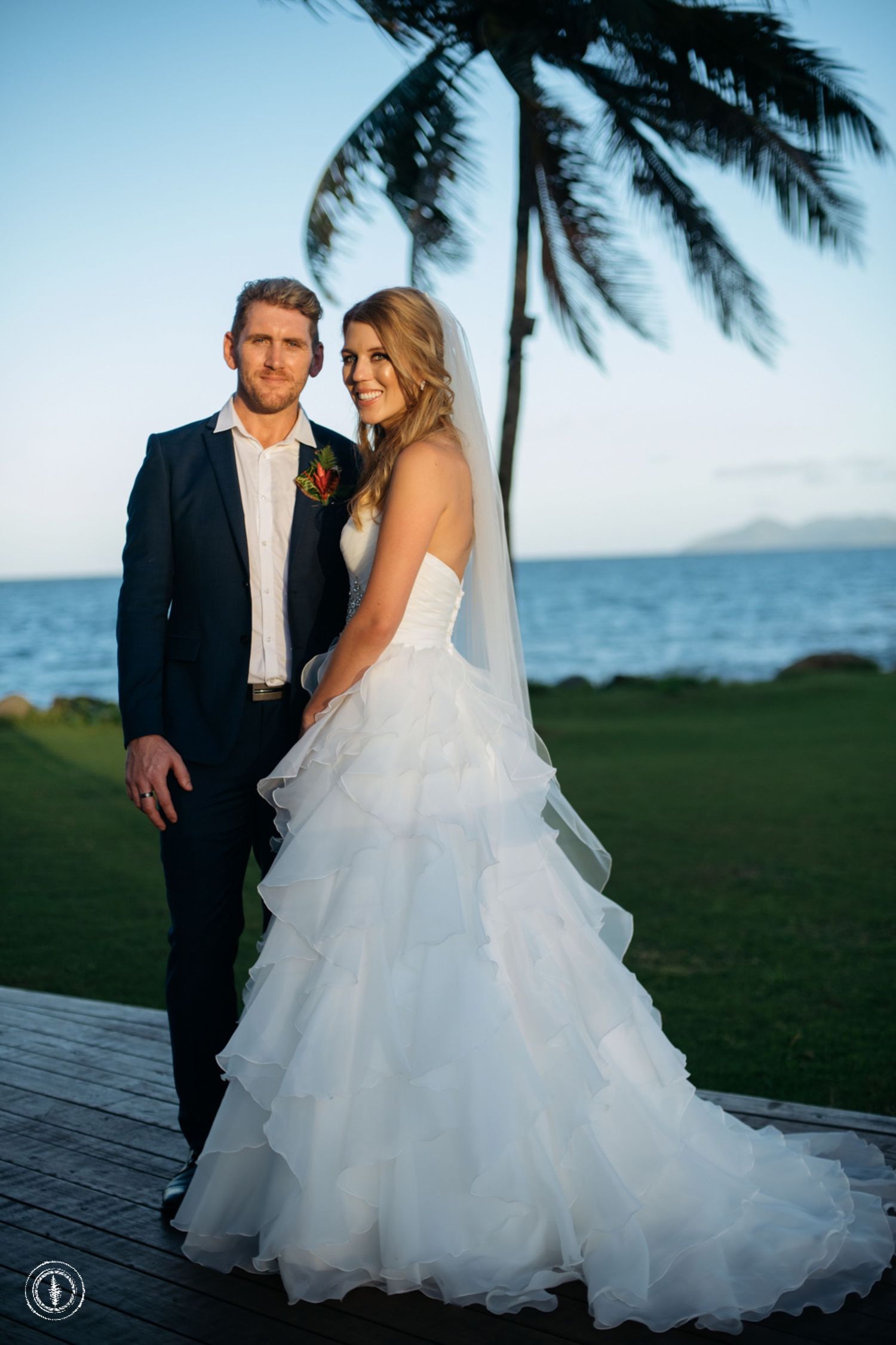 Elegant wedding couple poses on a lawn with palm trees and ocean views at sunset.