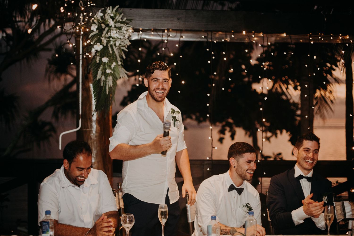 Groomsmen giving speeches at outdoor wedding reception with string lights.