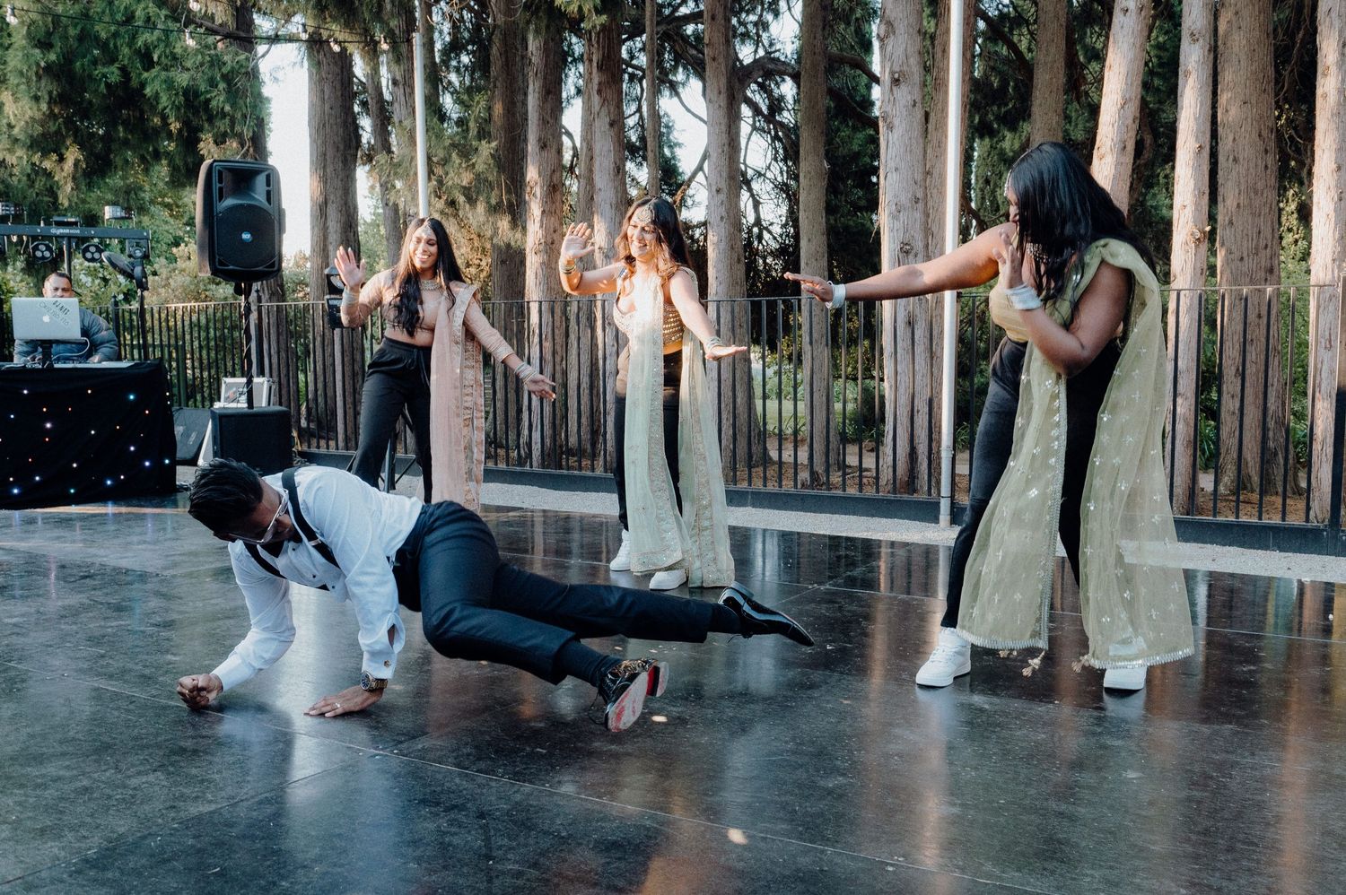 Wedding dancers performing energetic moves on an outdoor stage surrounded by trees.