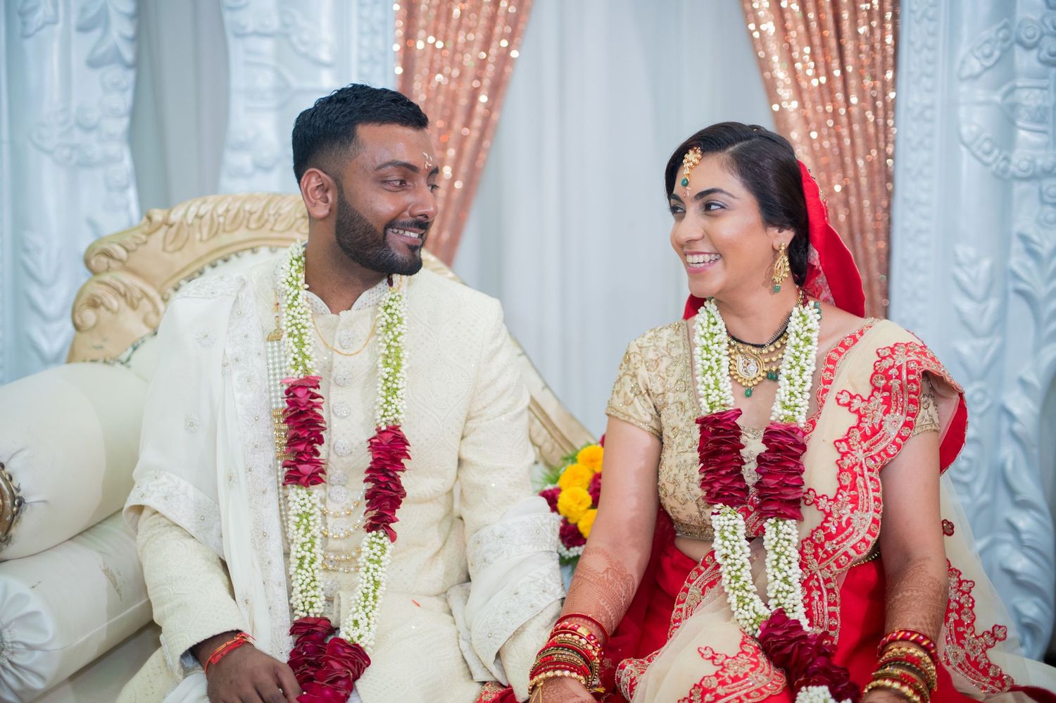 Newlyweds wearing traditional South Asian wedding attire with red garlands seated on an elegant white sofa.