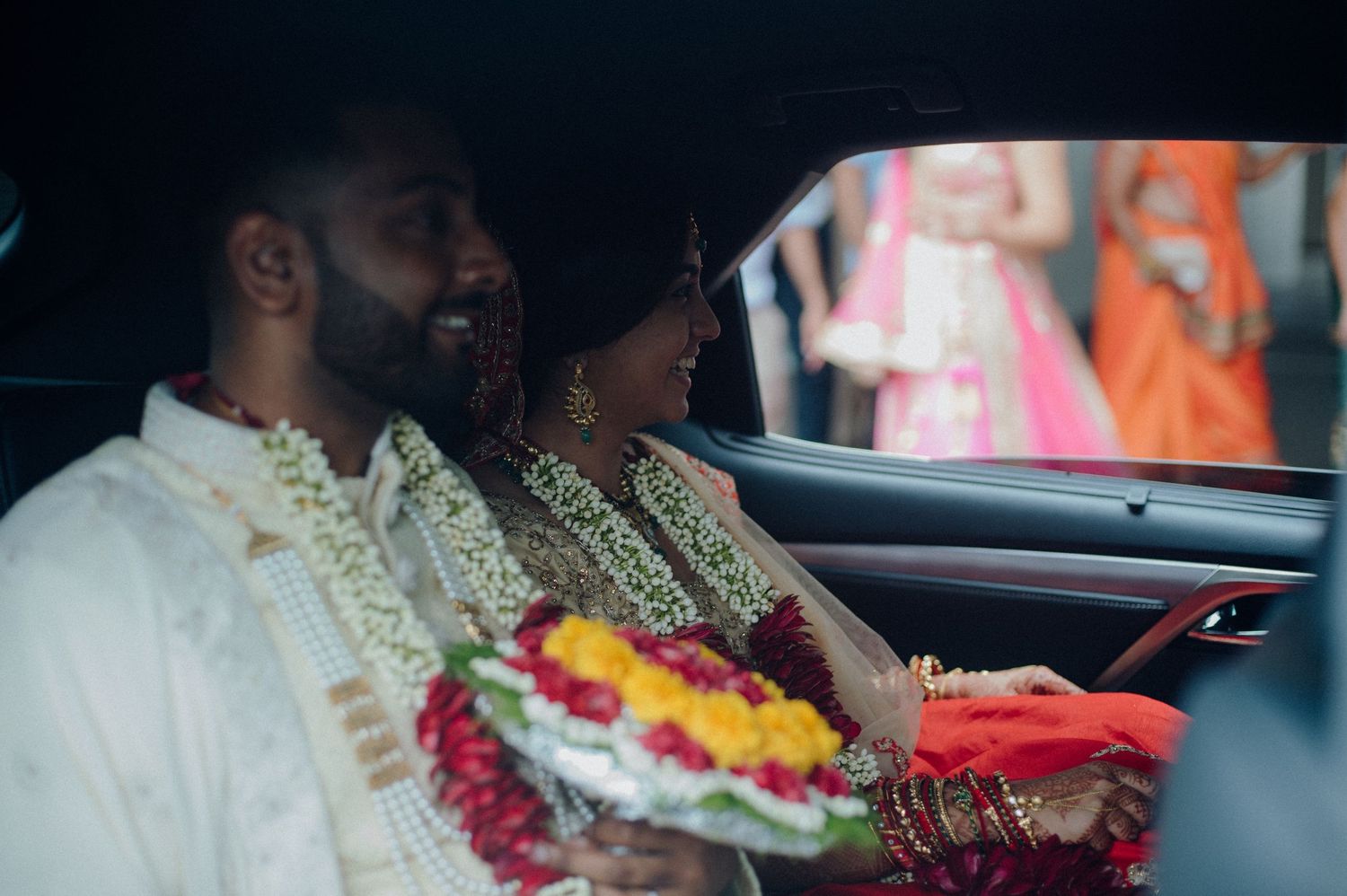 Couple in traditional wedding attire sitting in car with floral garlands.