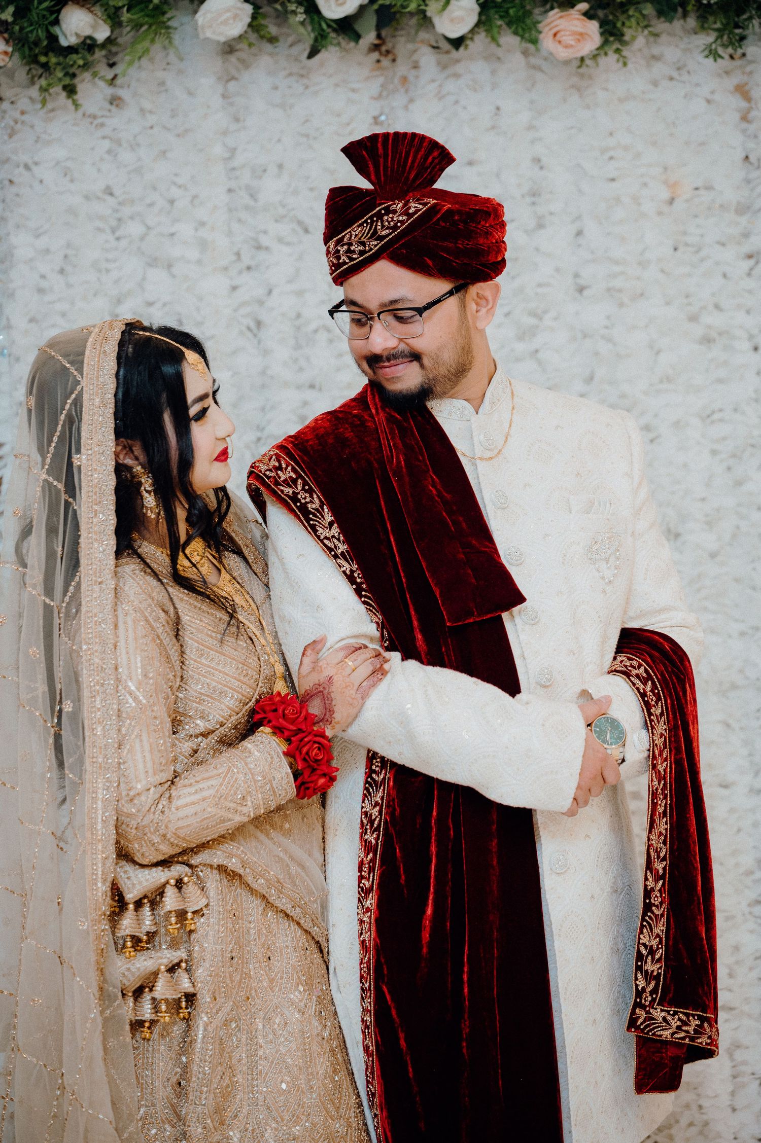 Couple in traditional wedding attire smiling against floral backdrop.