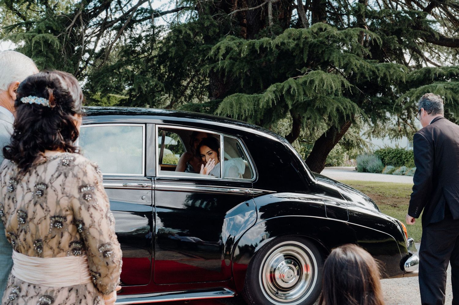 Wedding guests gather around vintage car as bride arrives at outdoor venue.