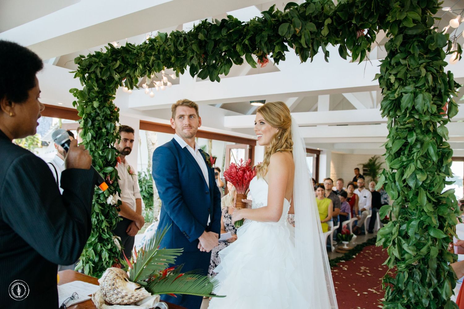 Wedding ceremony under lush green archway with red floral accents in an elegant indoor venue.