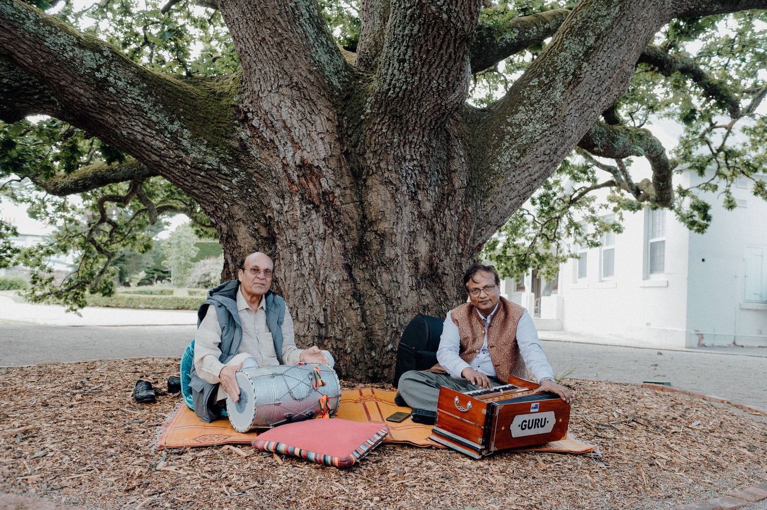 Two people relaxing on picnic blankets under a large oak tree with music equipment.