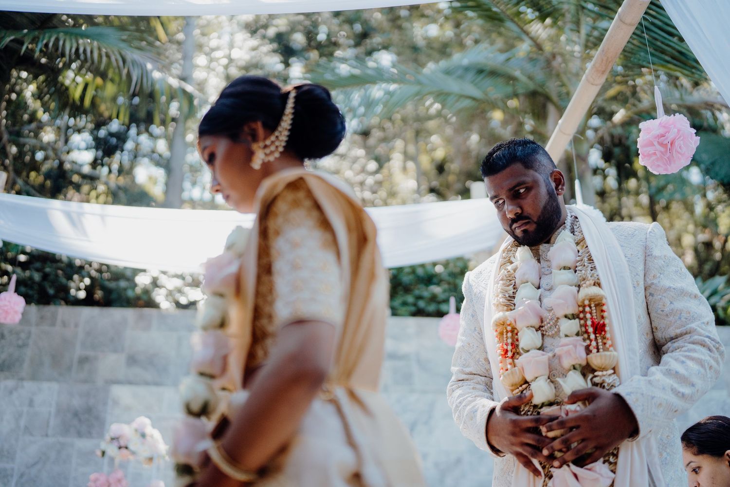 Indian groom in white sherwani with floral garland watches bride during outdoor Hindu wedding ceremony.