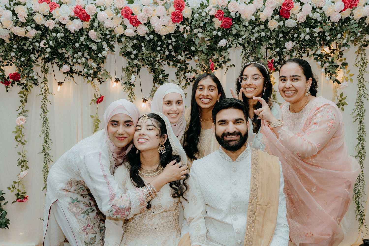 Group photo at wedding celebration with floral backdrop and people in coordinated white and pink outfits.