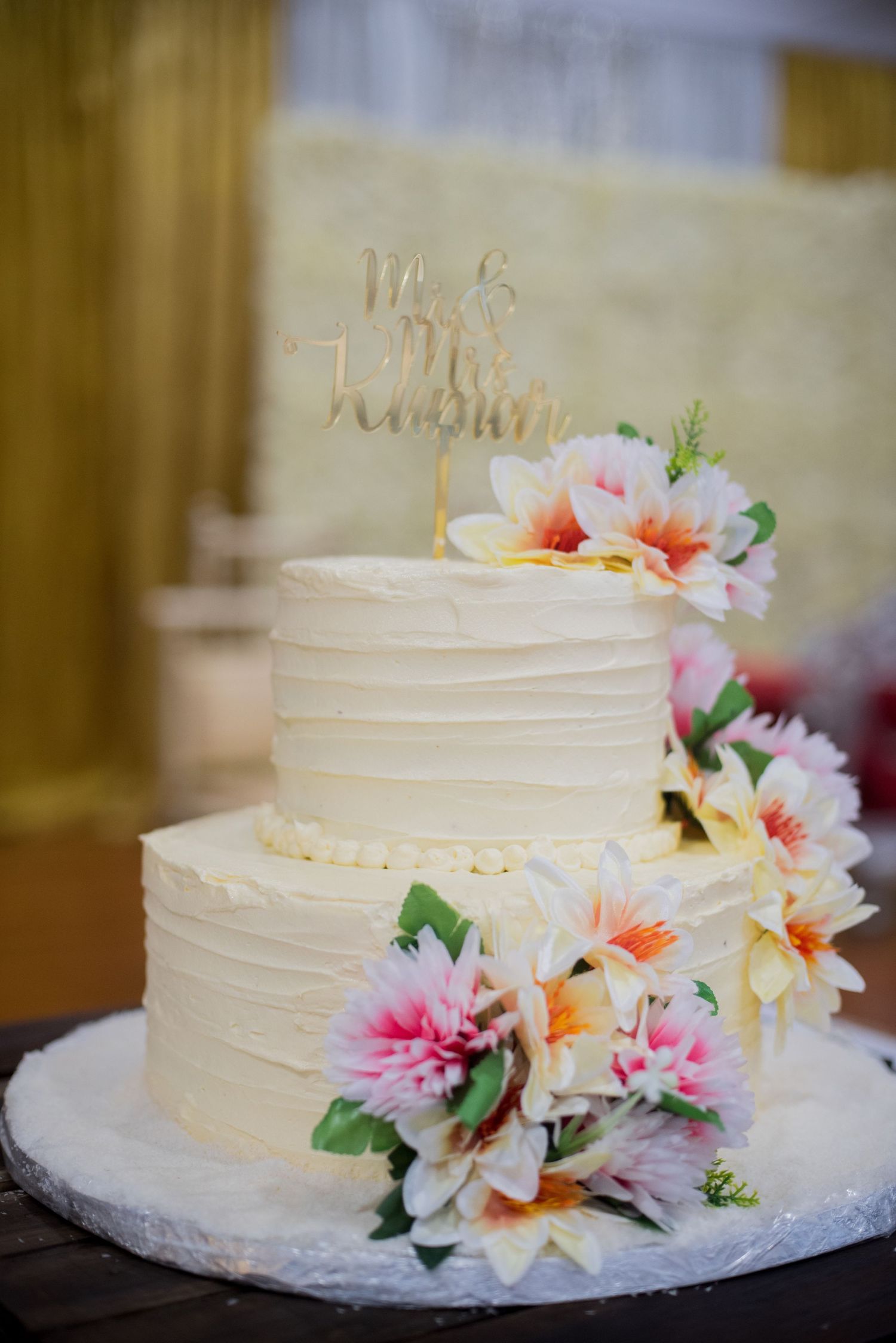A two-tier white wedding cake decorated with pink and peach flowers and a gold cake topper.