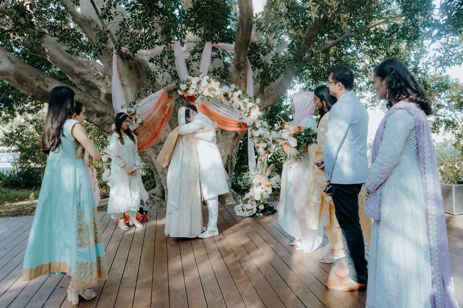 Wedding party and guests celebrate on wooden deck under trees with floral and fabric decorations.