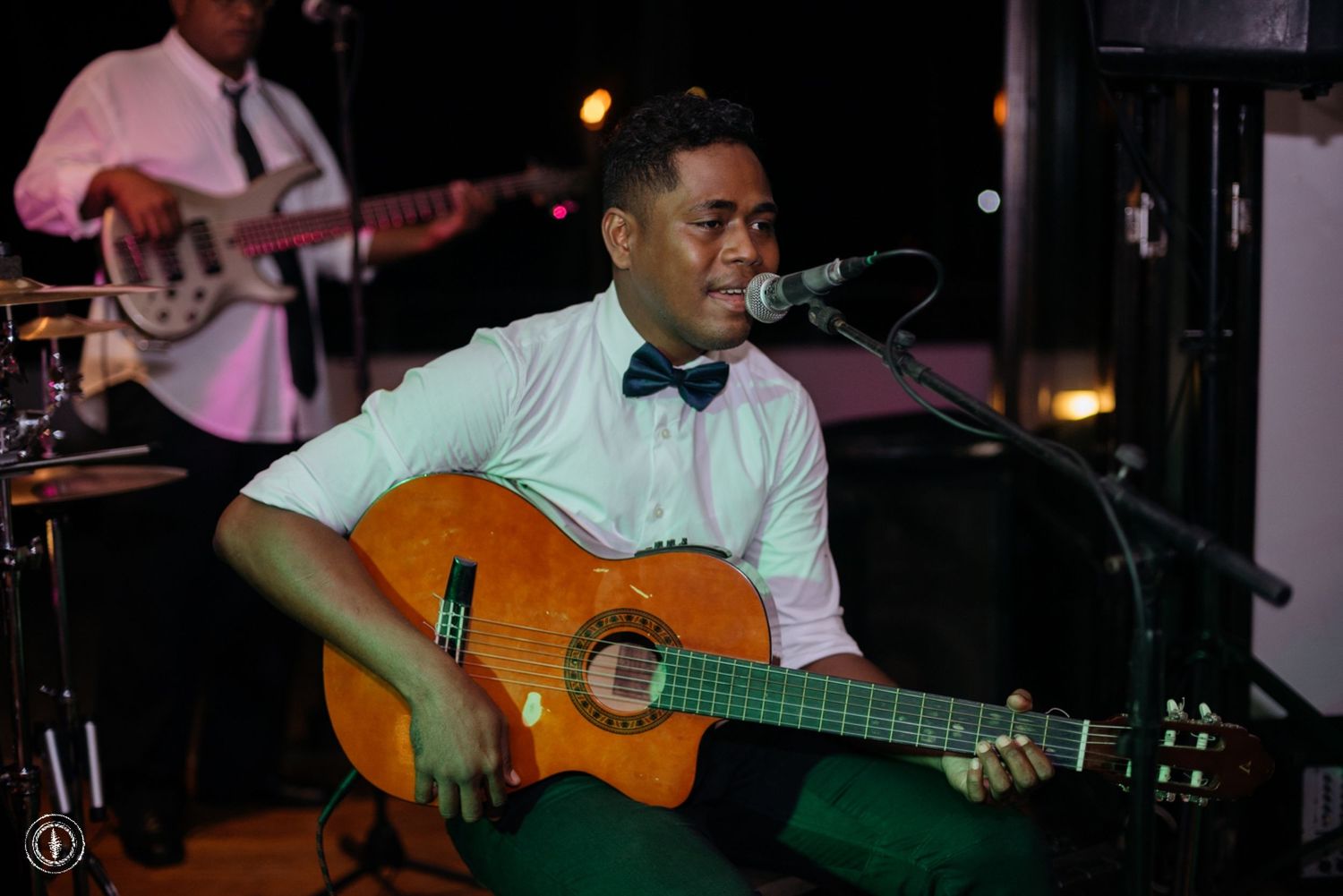 A musician plays an acoustic guitar while performing at an indoor venue with a bow tie and light colored shirt.