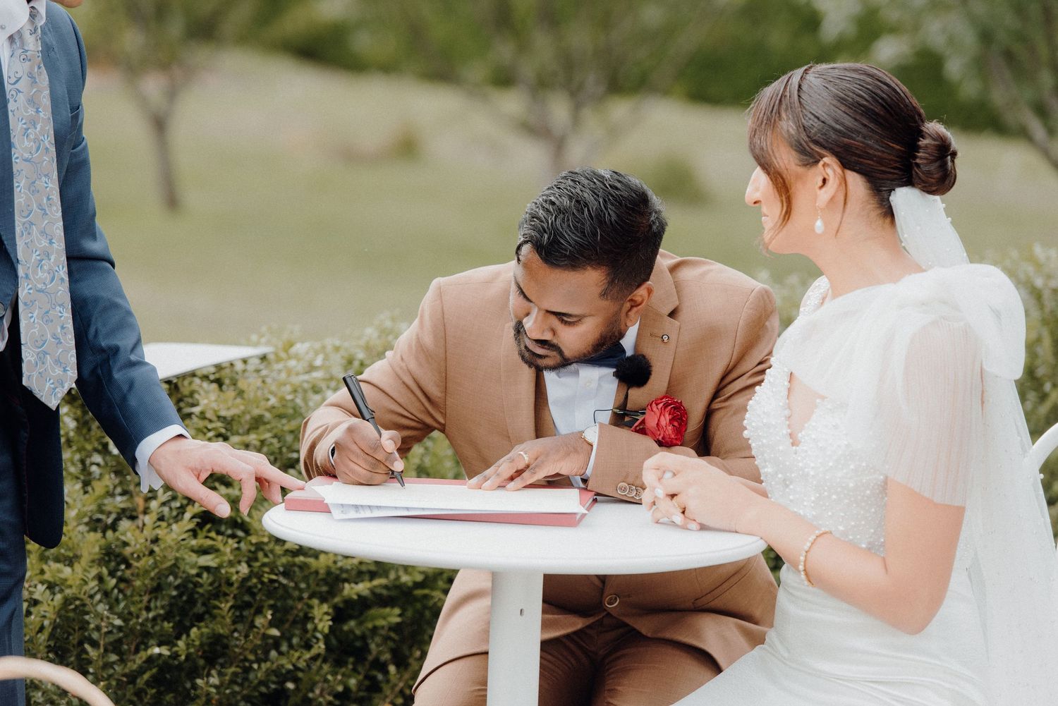 Wedding couple completing paperwork at a small white table during outdoor ceremony.