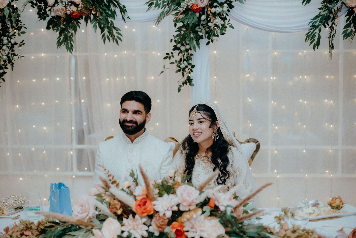 Reception setting with floral decor, twinkle lights, and couple at head table.