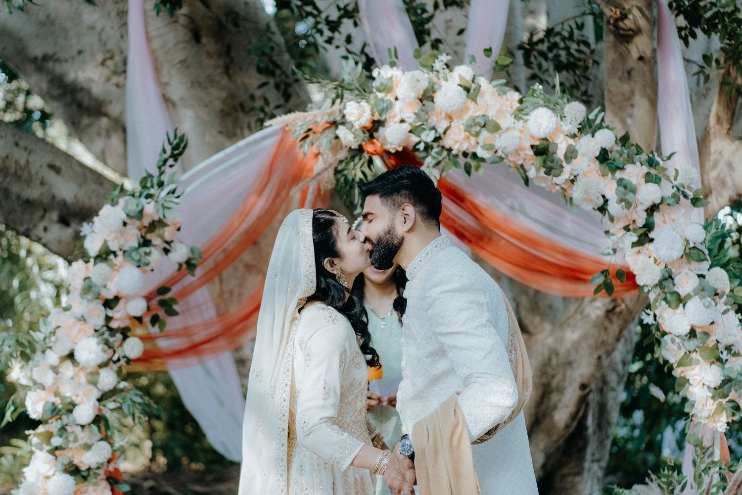 Intimate moment between newlyweds under a garden arch decorated with flowers and coral fabric.
