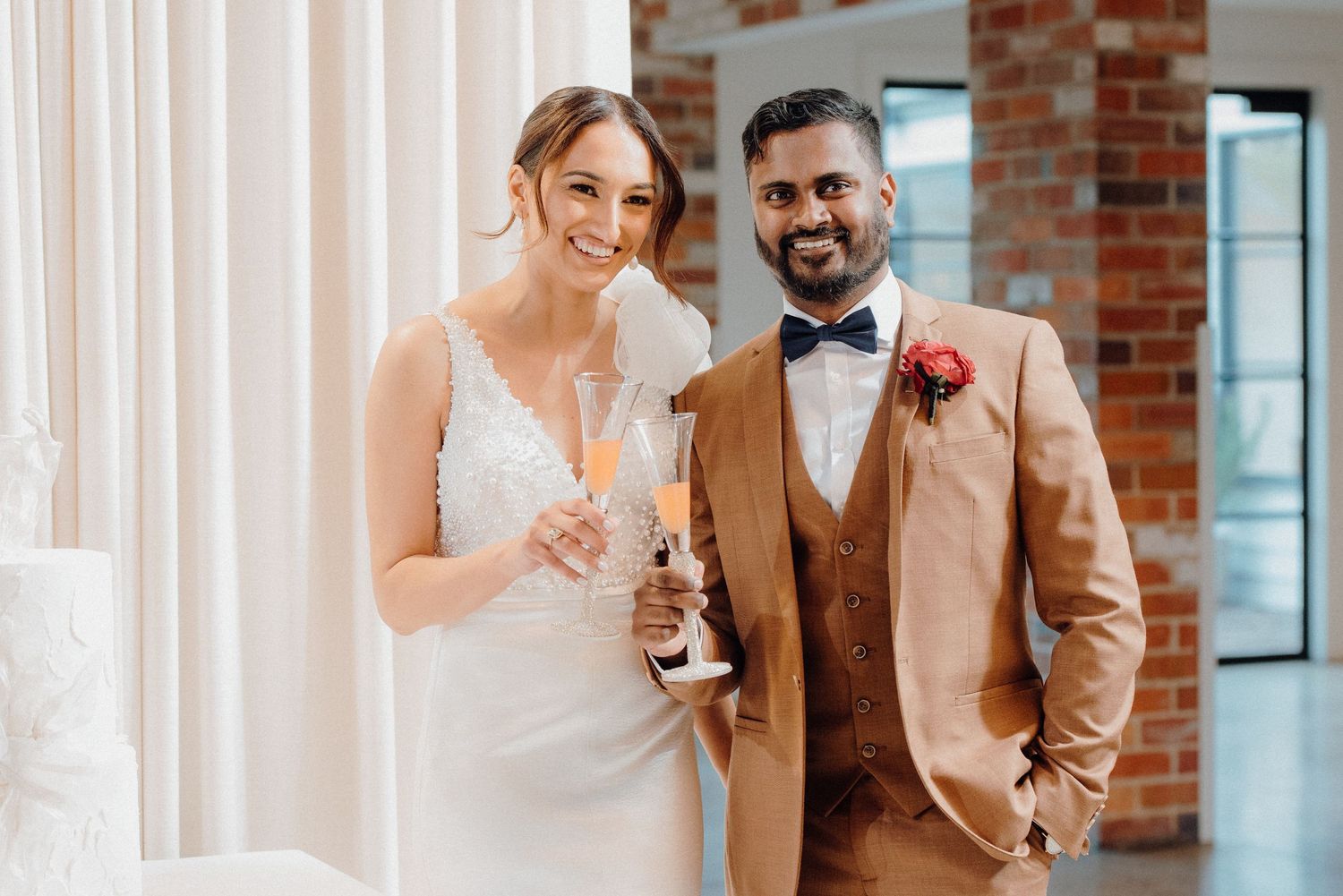 Couple in wedding attire pose with champagne glasses against white curtain backdrop.
