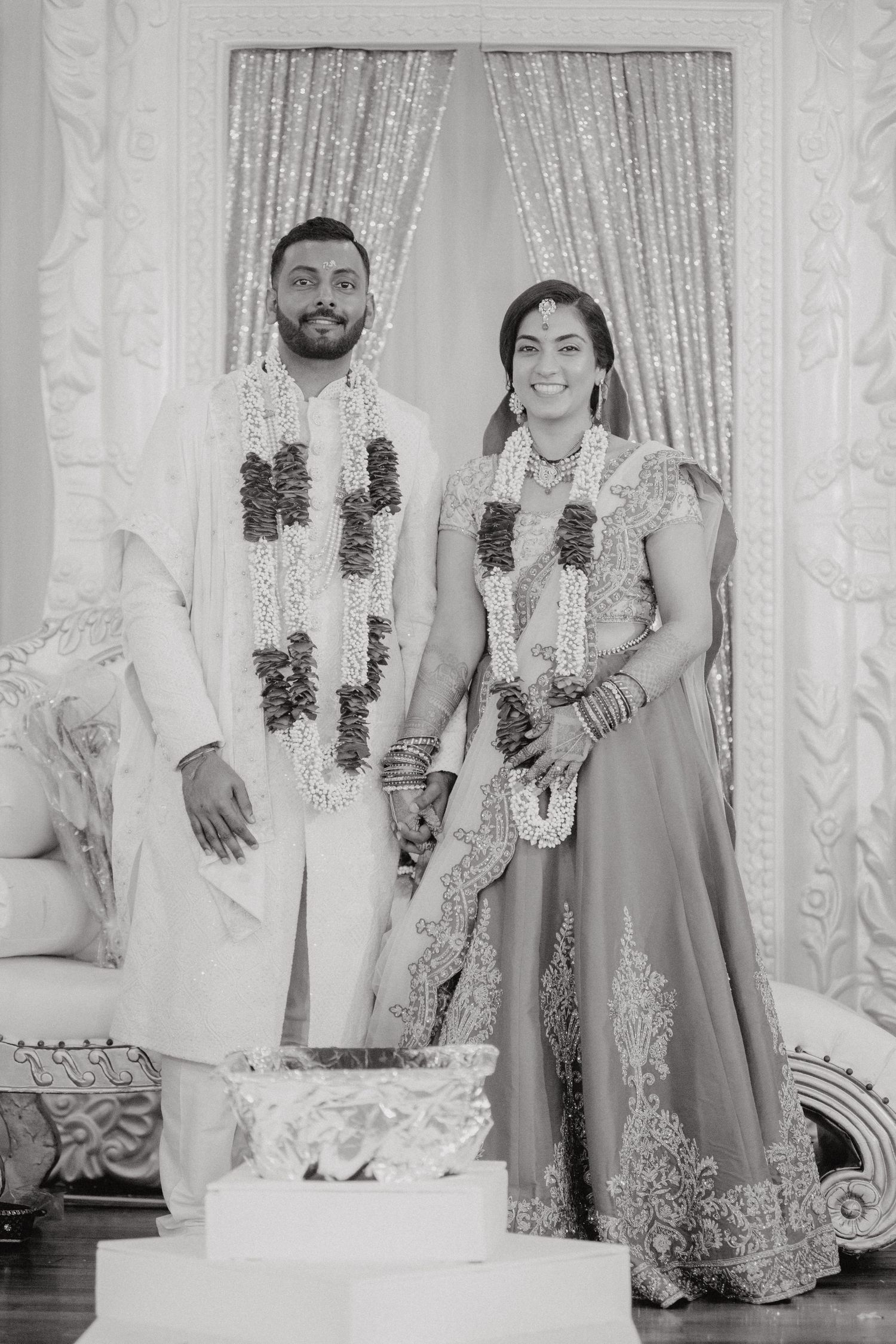 A couple in traditional Indian wedding attire stands together against a silver backdrop.