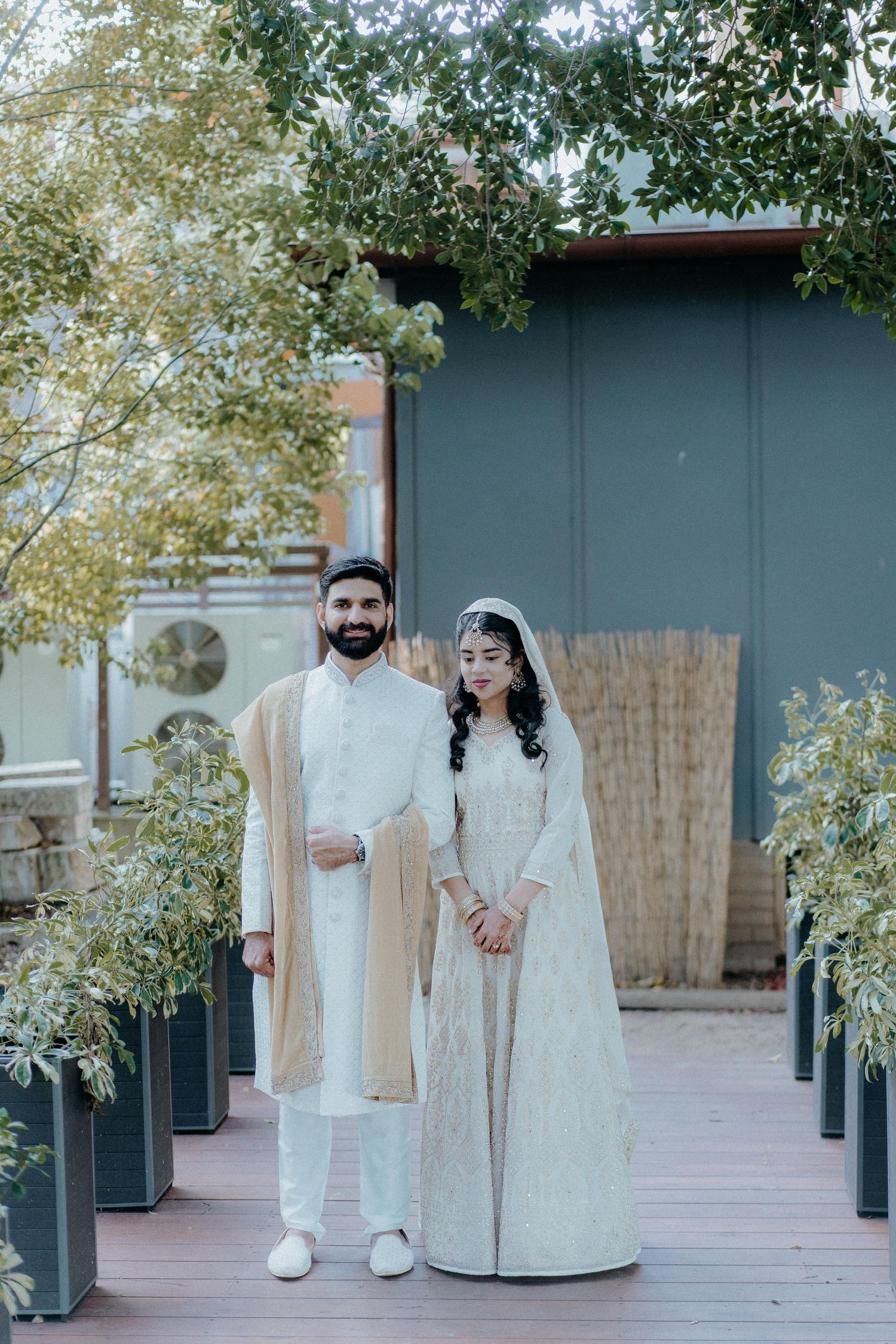 A couple in traditional white and cream wedding attire poses on a wooden deck surrounded by potted greenery.