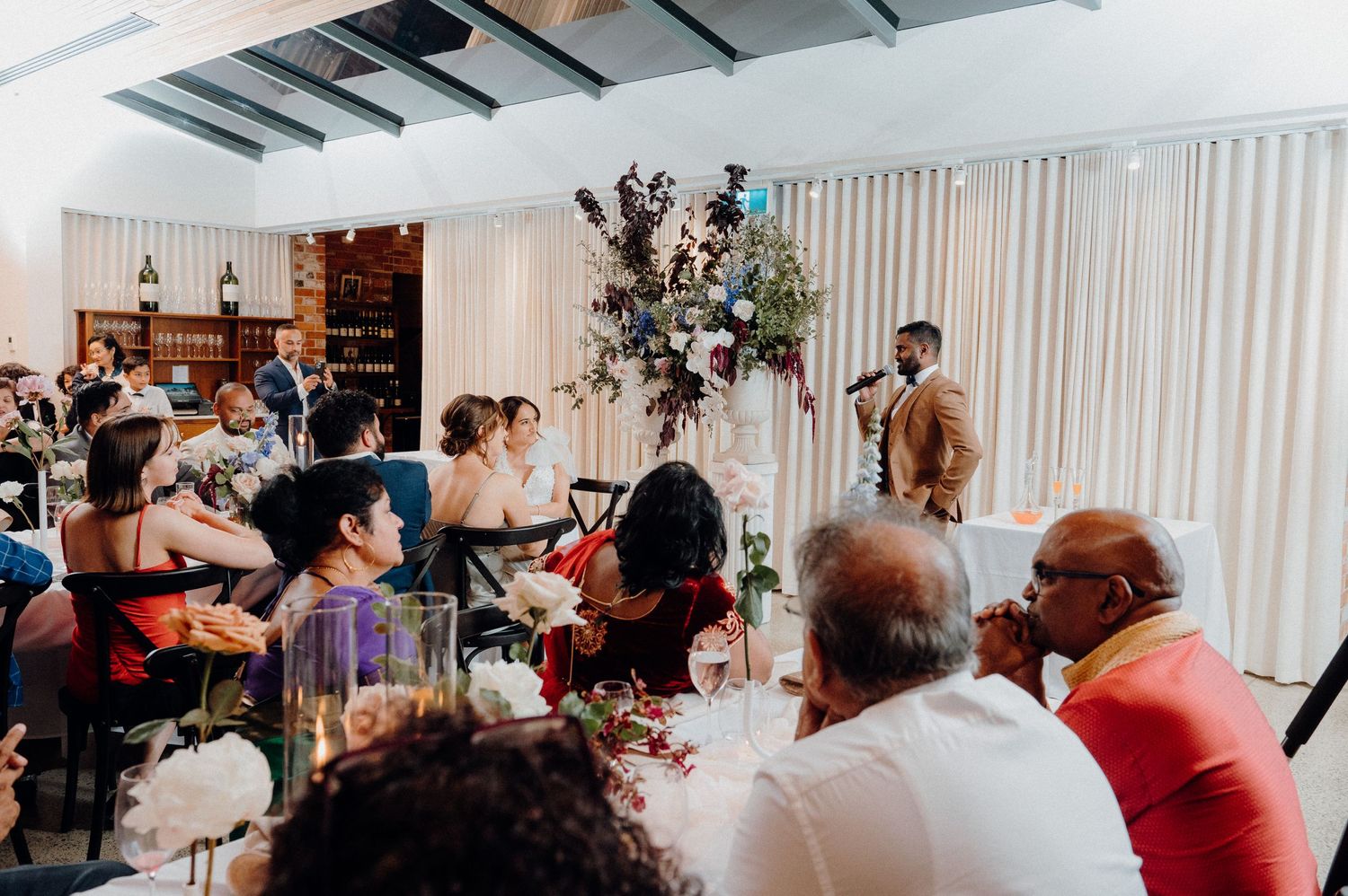 Wedding reception guests gathered around decorated tables with floral centerpieces.