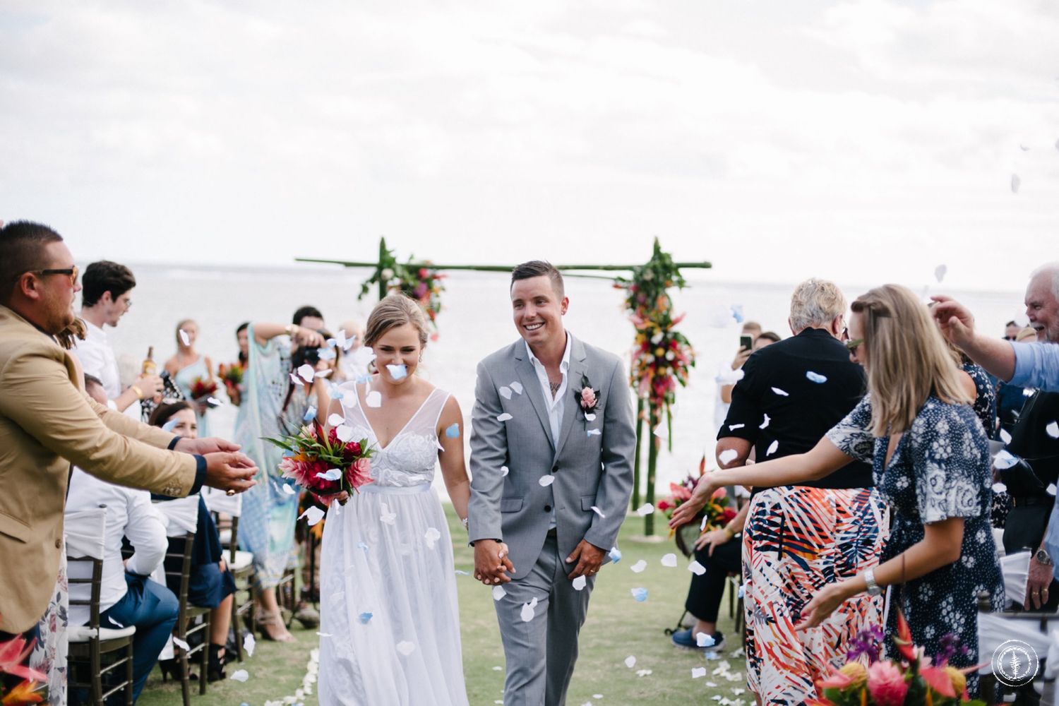 Newlyweds walk down the aisle as guests celebrate at an outdoor oceanside wedding.