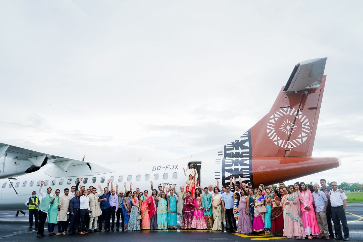 Large group photo of wedding party in colorful attire posed in front of commercial aircraft.