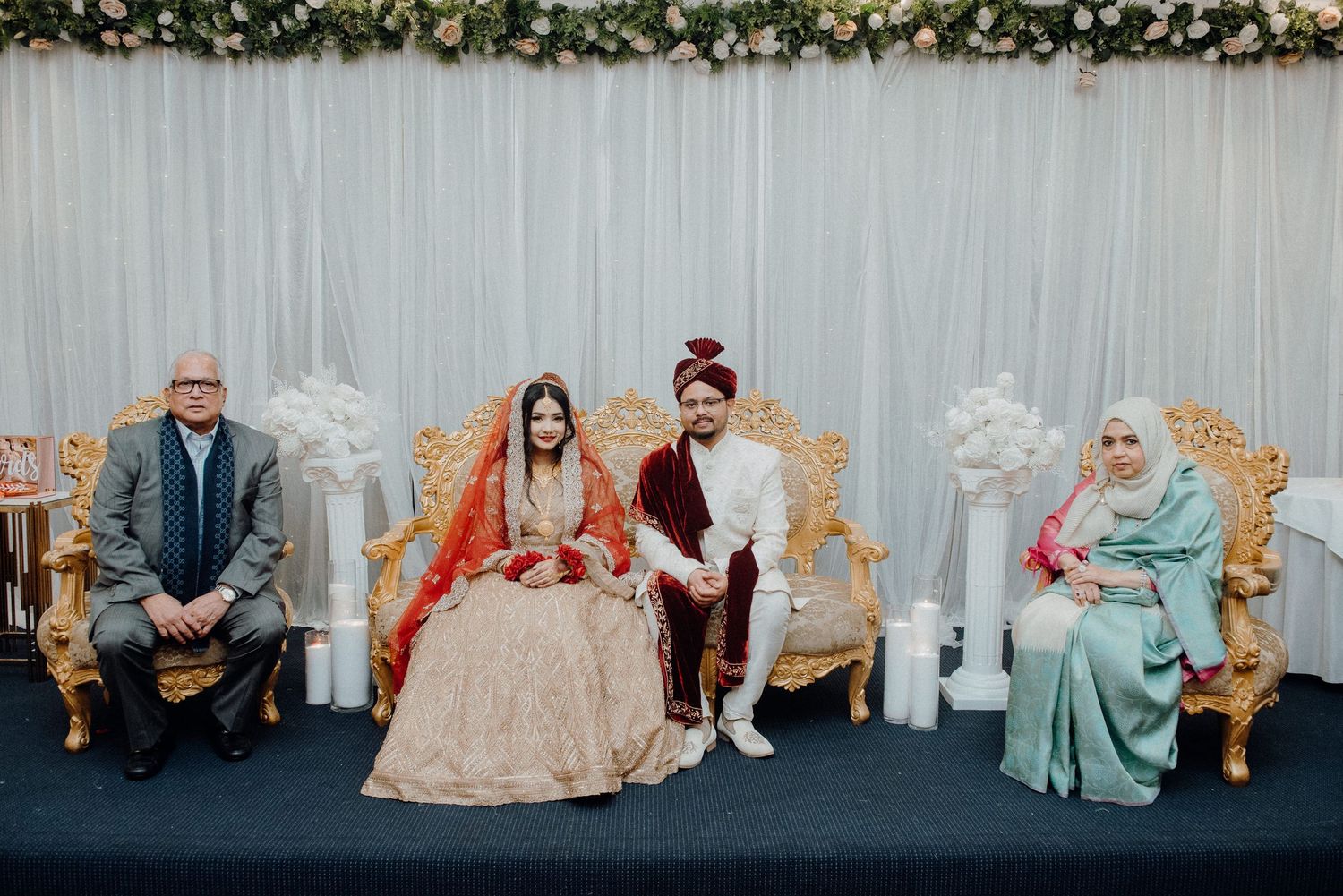 Wedding party seated on ornate chairs with white decorations and floral garland.