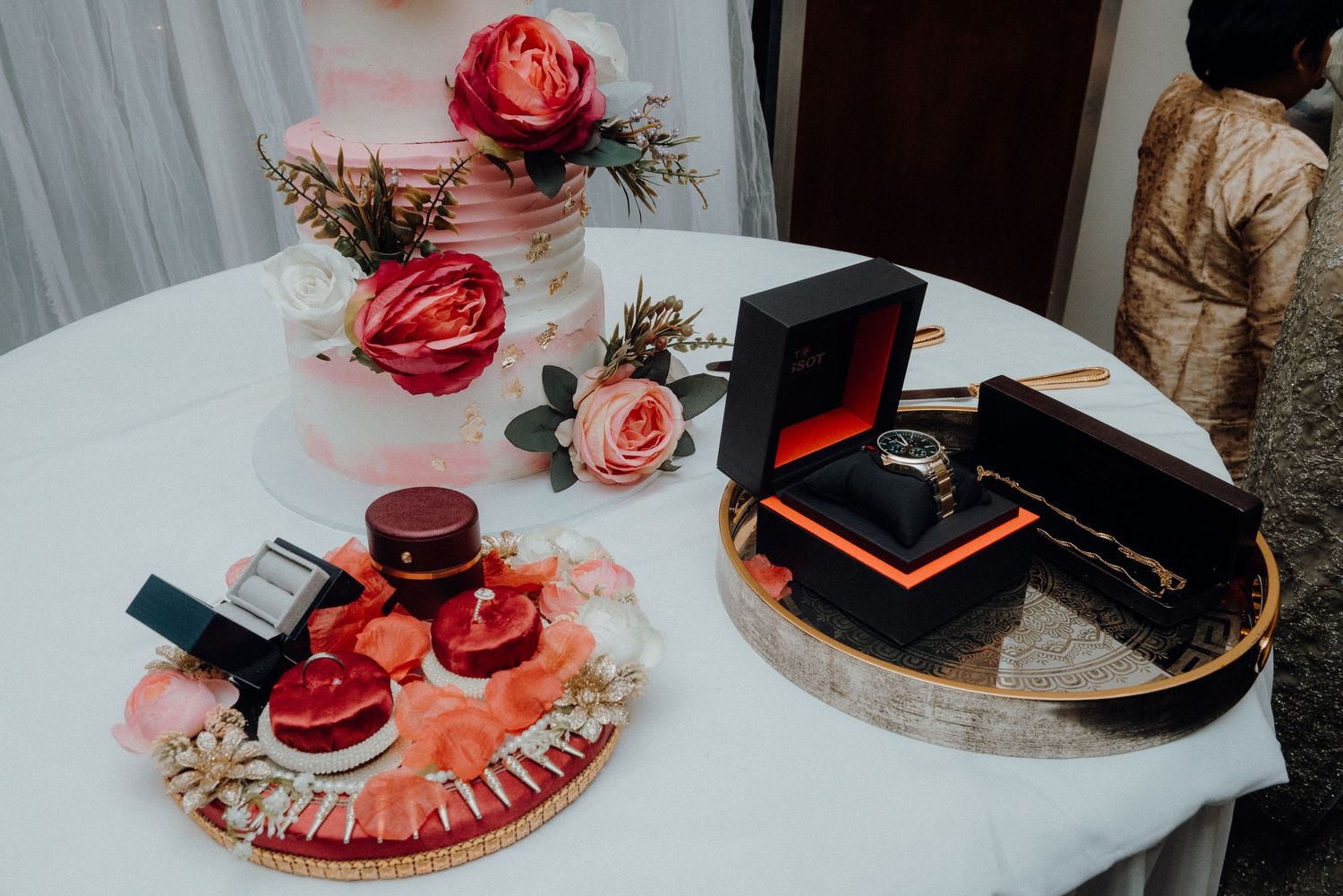 Elegant wedding decor with red roses and jewelry boxes displayed on white tables.