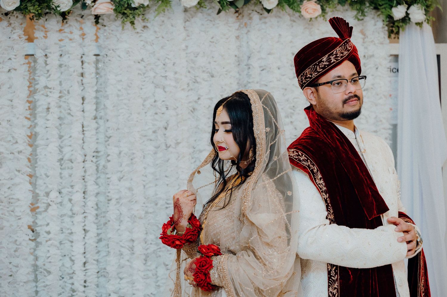 Couple in traditional wedding attire pose back to back against floral backdrop.