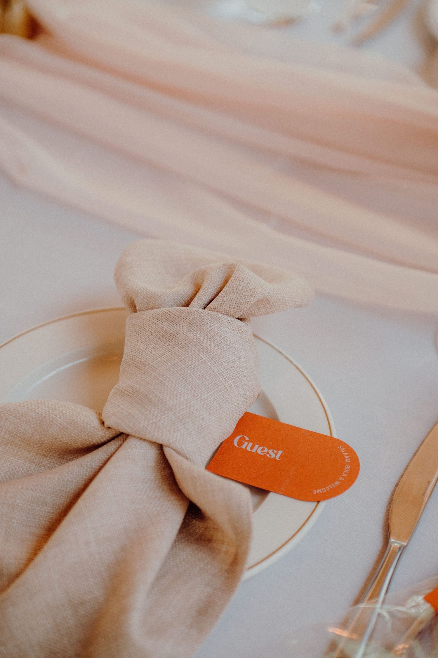 Close-up of beige napkin folded with orange guest card on white plate.