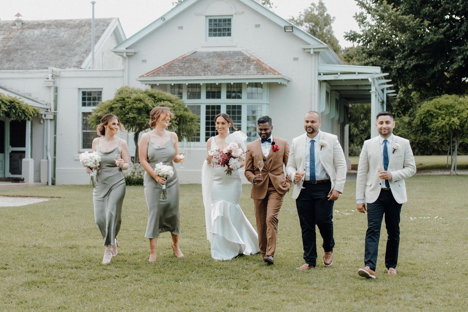 Wedding party walking and laughing together on grass near white house.