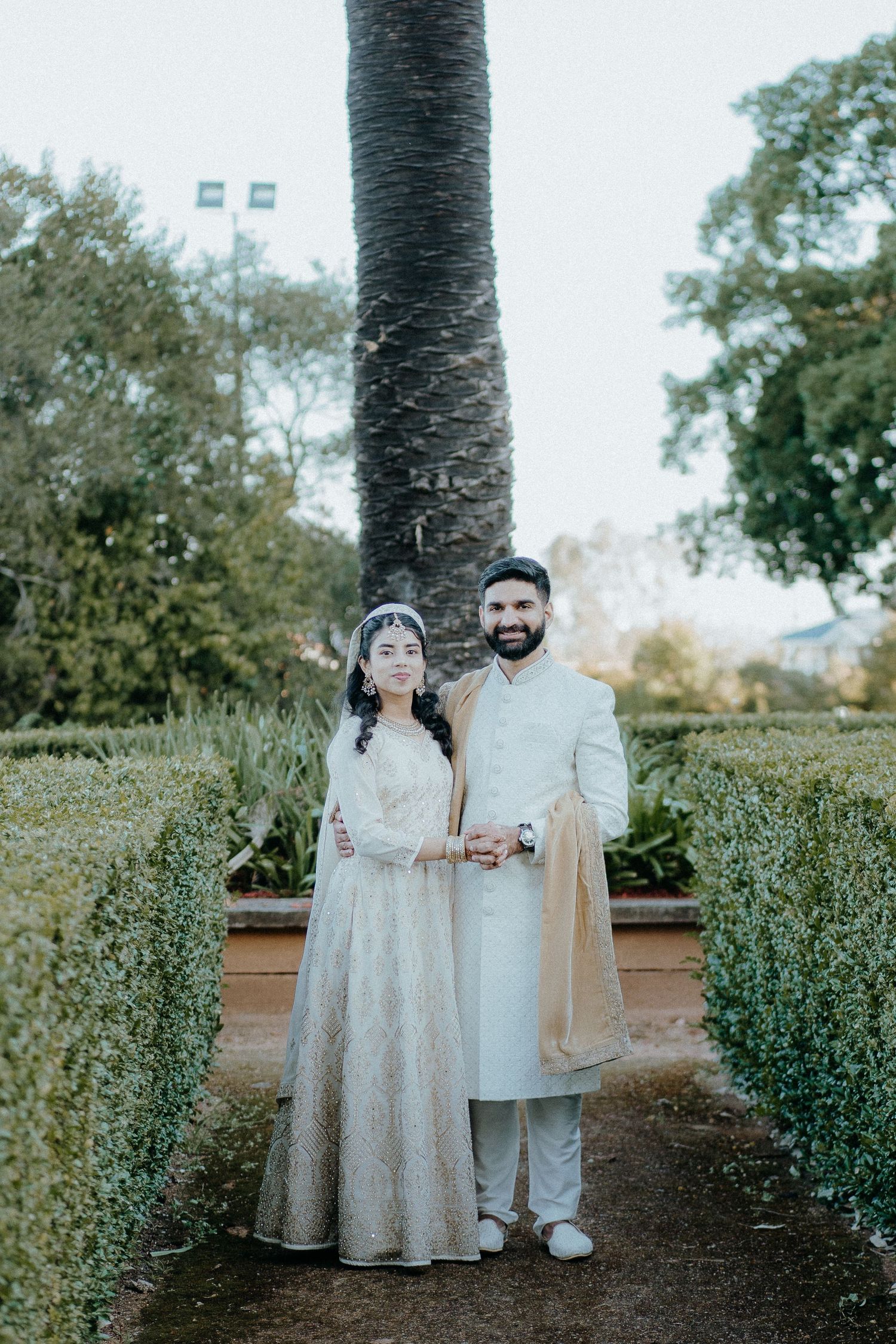 Couple in traditional wedding attire posing in garden setting with palm tree backdrop.