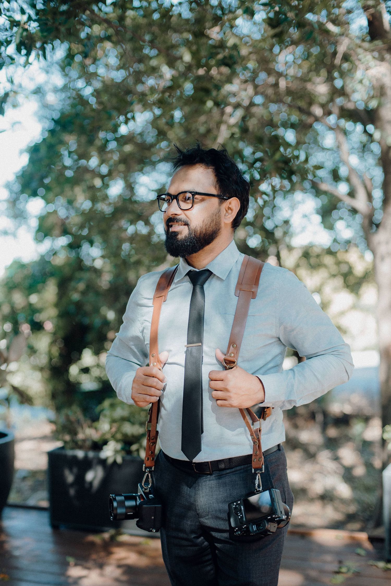 Professional wedding photographer wearing glasses and dual camera straps standing outdoors among trees.