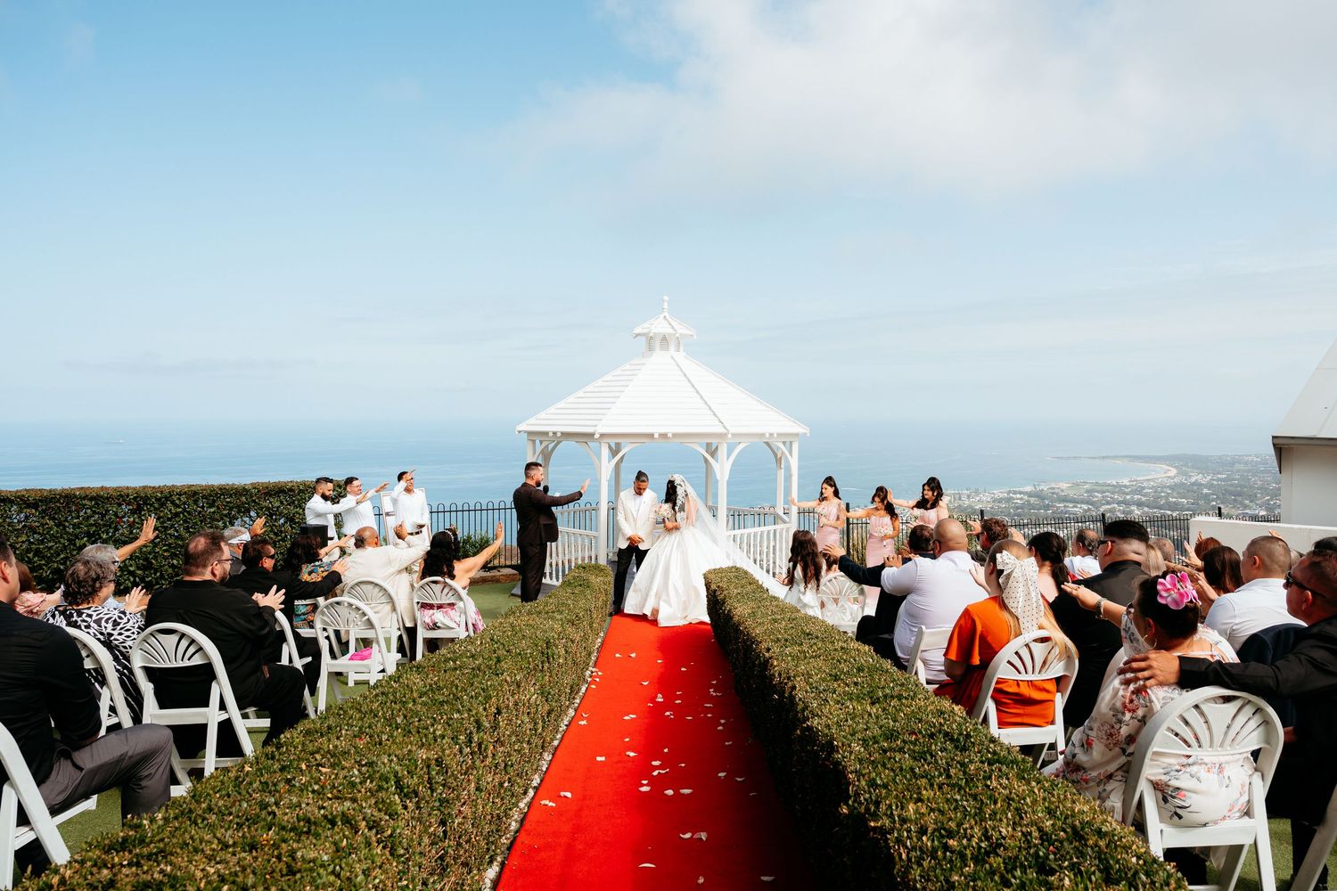 Elegant outdoor wedding ceremony at white gazebo overlooking ocean with red petal aisle.