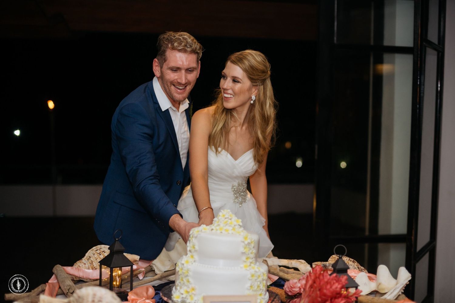 Couple cuts their elegant white wedding cake together at evening reception.